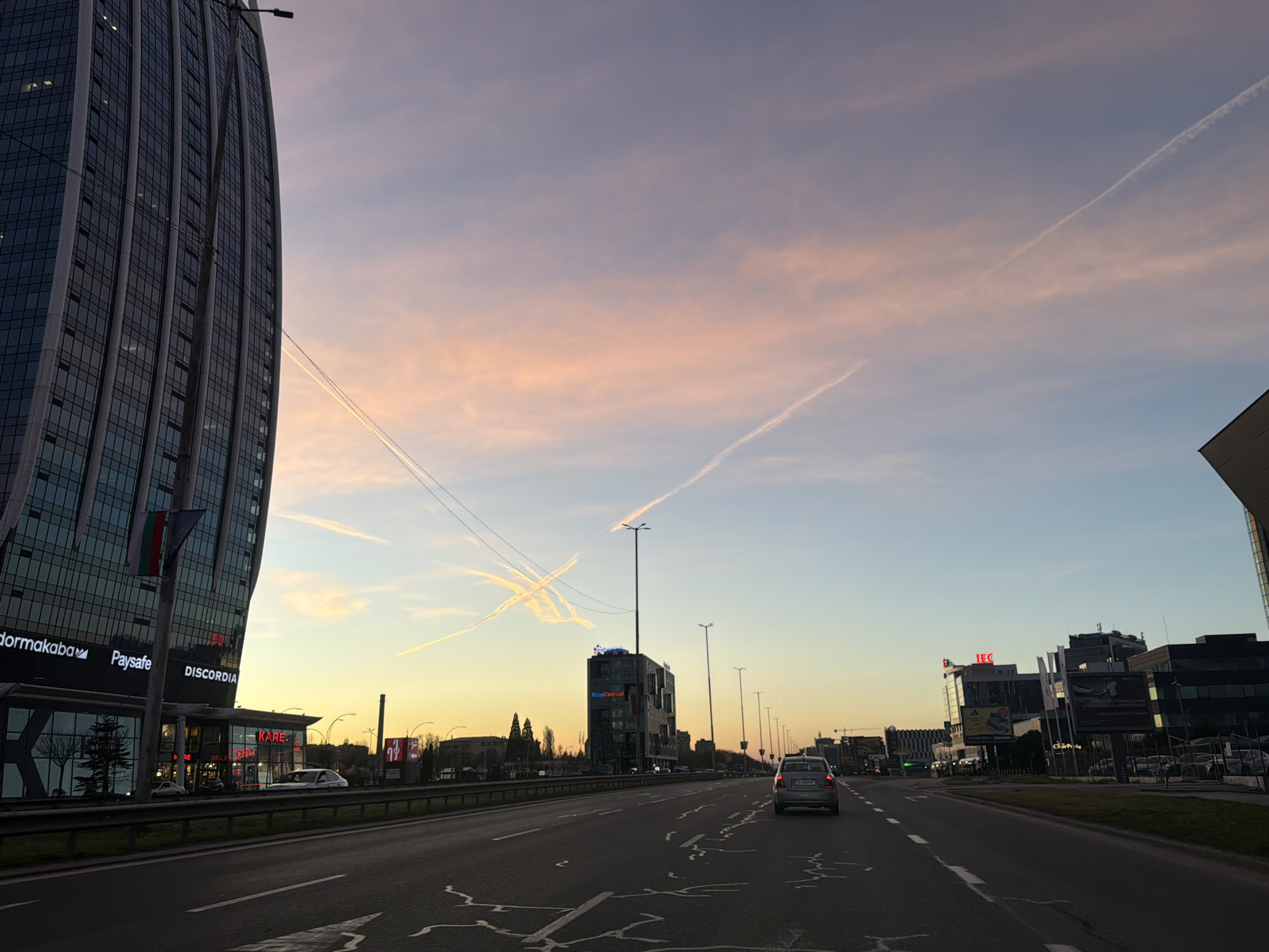 The image depicts a multilane highway in Sofia, Bulgaria, lined with modern office buildings and illuminated by streetlights. A gray car is visible in the middle of the right-hand lane, moving away from the viewer.
In the background, various commercial and office buildings are present, including ones with signage for "dormakaba," "Paysafe," "DISCORDIA," "KARE," "RingCentral," and "IEC." A tall, curved glass building is prominently featured on the left, adorned with flags.
The sky is a gradient of colors, from a deep orange near the horizon to light pink and blue higher up. Vapor trails are visible crisscrossing the sky. Road markings are present on the asphalt, indicating lane divisions and directional arrows. The time of day appears to be sunset or sunrise.
