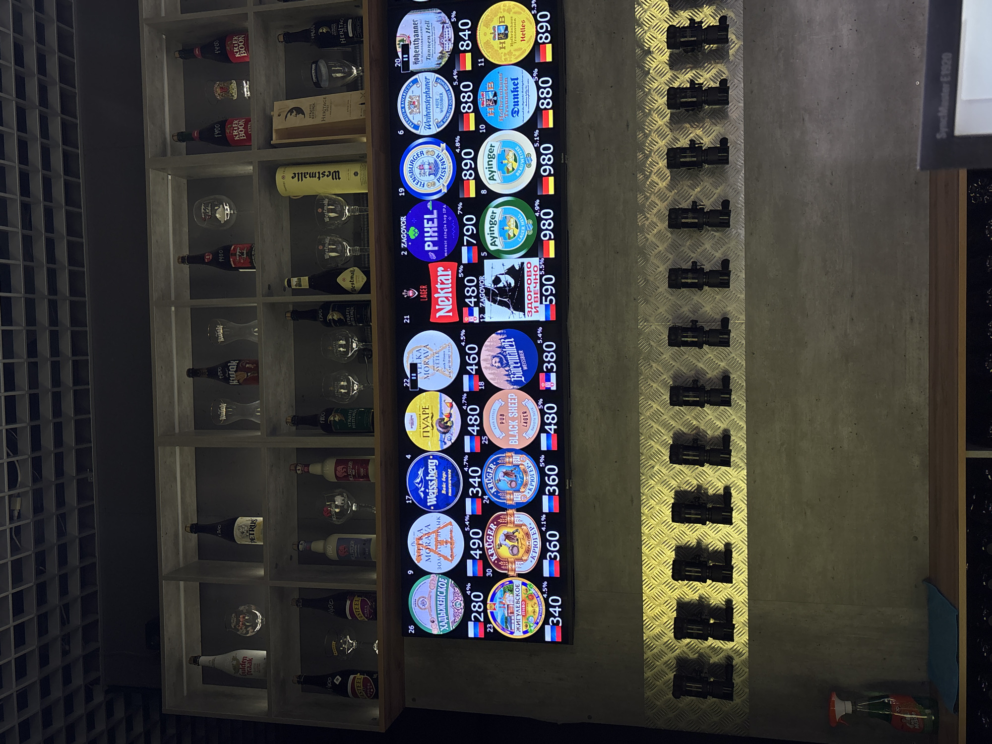The image shows a display of various beers on tap in what appears to be a bar or restaurant. The display features a digital screen showcasing beer logos, names, and prices, positioned next to a shelving unit containing various bottled beers and glassware. Below the digital display is a section with beer taps.
The beers listed on the screen include both local and international brands, with prices and alcohol content (%) shown beside each brand. The beers include the following brands and names: "VELKA MORAVA," "ХАДЫЖЕНСКОЕ," "ЖИГУЛЕВСКОЕ," "PUARE," "KRUGER," "PUB BLACK SHEEP," "VELKA MORAVA," "NEKTAR," "BärentaleR," "ZAGOVOR," "PIXEL," "Ayinger," "FLENSBURGER," "Weihenstephaner," "Hofbräuhaus," and "Hohenthanner."
The bottles on the shelf include brands such as "ASTEEL," "PETRUS," "KRIEK BOON," and "Westmalle."
The digital screen also features "SyncMaster E1920," probably indicating the brand of the display screen.
The location may be in Vnukovo, Russia, since some brands on the list are commonly found in Russia.