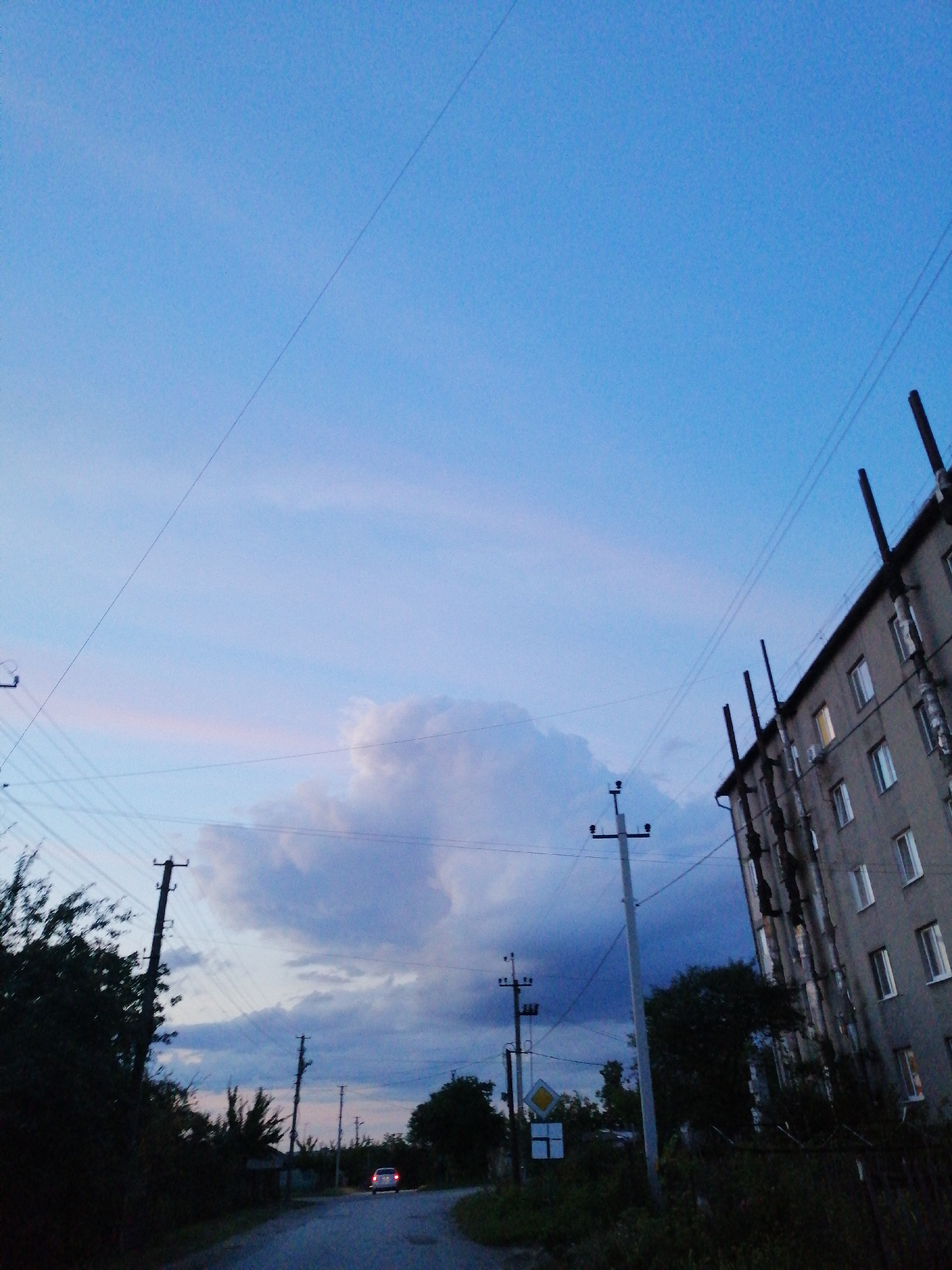 An outdoor, low-angle, wide shot captures a suburban street scene at dusk. The sky, a gradient of pale blue to muted purple, features wispy clouds. A large, multi-story apartment building, constructed of grey concrete, dominates the right side of the frame. Its facade is marked by numerous windows, some illuminated from within, and vertical pipes run down its side. A telephone pole stands in front of the building. Several other telephone poles and a dense cluster of dark trees line the left side of the image, partially obscuring a road that curves into the distance. The road surface is asphalt, and in the far distance, a single white car with its taillights illuminated is visible, driving away from the viewer. A yellow diamond-shaped traffic sign is present on the left side of the road.