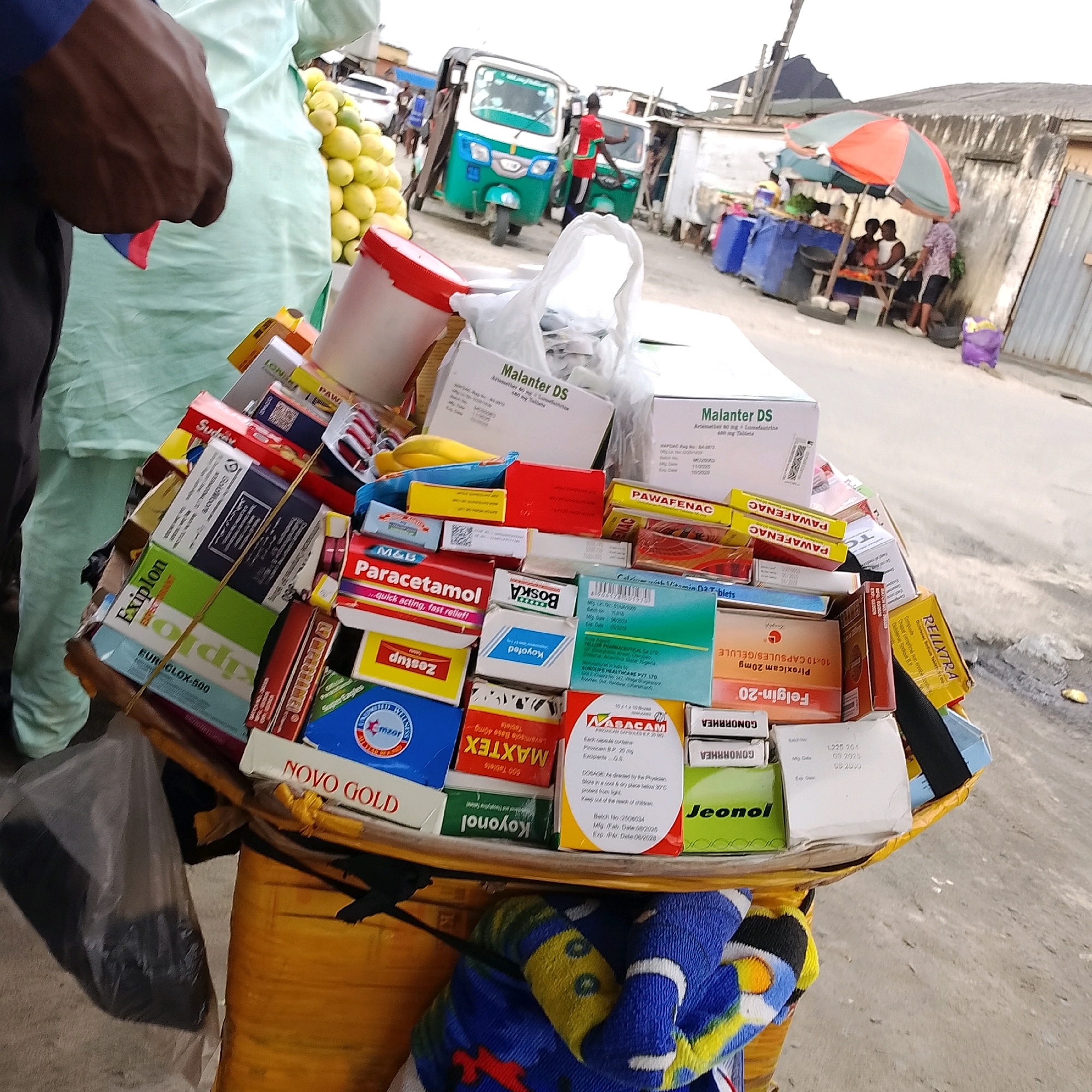 The image depicts a street vendor in Ado, Nigeria, with a cart overflowing with various medications. The cart is laden with boxes of pharmaceuticals, including brands like Malanter DS, Paracetamol, Exiplon, PawaFenac, Felgin-20, and Jeonol. Some boxes are upright, while others are stacked horizontally, creating a dense collection of products. In the foreground, a person's arm and shoulder are visible on the left, clad in a dark sleeve and blue fabric. The vendor is standing behind the cart, though not fully visible. The background shows a busy street scene with a blue and white tricycle, a red and green umbrella providing shade over a stall with people gathered, and other buildings and market stalls typical of an urban Nigerian environment. The ground is paved and appears slightly dusty.