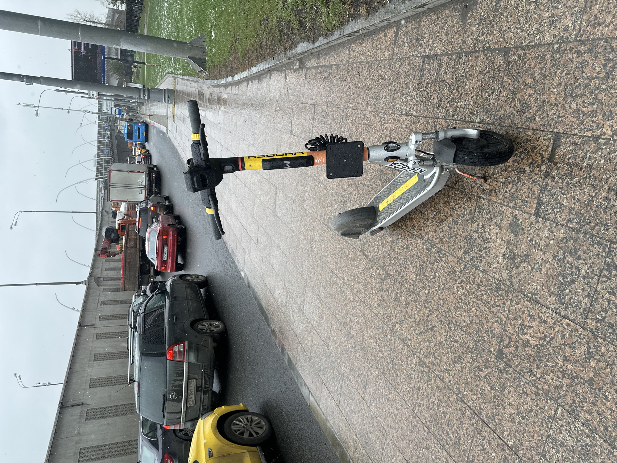 The image shows a Woosh electric scooter parked on a paved sidewalk in Moscow, Russia. The scooter is silver and yellow with the registration number KX 762A visible on the frame. It is parked near a road with heavy traffic. Cars are lined up in a queue and include a yellow car, a grey Opel Astra, and a red car. A truck and other vehicles are visible further down the road. The environment appears to be urban and potentially industrial, given the presence of a bridge or overpass with noise barriers. The weather is overcast and possibly rainy or snowy, indicated by the wet pavement and what appears to be some snow or ice on the grass in the background.