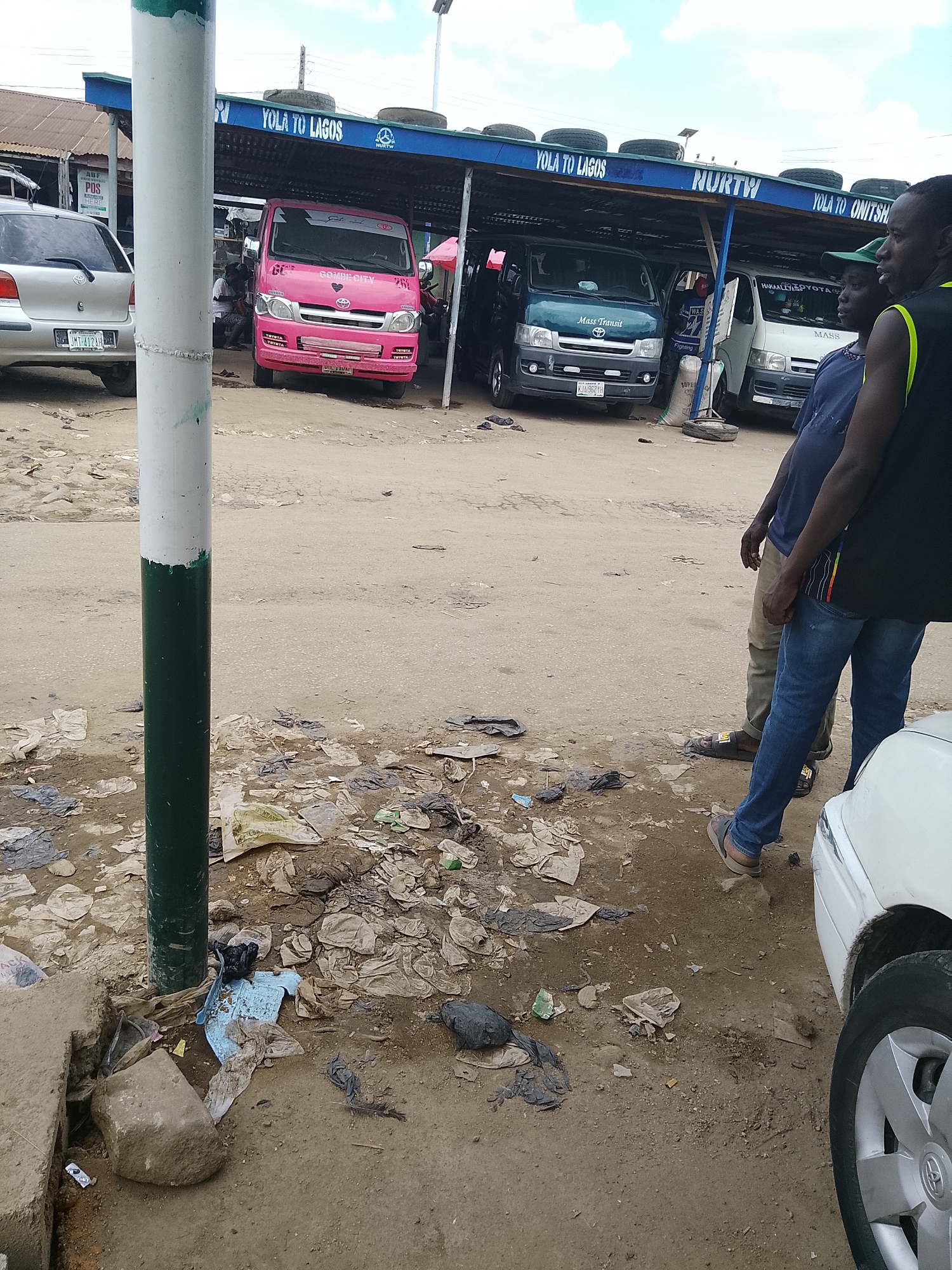 The image depicts an outdoor scene in what appears to be a commercial area, likely a transportation hub or a roadside market, in Wauro Leggal, Nigeria. In the foreground, a substantial white and green painted pole stands vertically, partially obscuring the view. The ground is unpaved and littered with a considerable amount of refuse, including plastic bags, debris, and some discarded tires.
Several vehicles are visible. To the left, a silver Toyota sedan is parked, its rear visible, displaying a license plate. Further into the scene, under a covered structure, a bright pink minibus with "GOMBECITY" written on its side is parked. To its right, a dark green minibus is parked, with "Mass Transit" written on its front. Next to that, a white minibus is also parked. The covered structure has signs indicating "YOLA TO LAGOS" and "YOLA TO ONITSHA".
Three individuals are present. Two men are standing in the right foreground, facing towards the vehicles and the covered structure. One man is wearing a blue t-shirt and jeans, while the other is wearing a black t-shirt and jeans. Their stances suggest they might be waiting or observing. A third person, partially visible in the pink minibus, appears to be seated inside.
The overall environment suggests a bustling, possibly informal, transportation terminal, characterized by the presence of commercial vehicles and a lack of meticulous sanitation. The sky overhead is partly cloudy.
