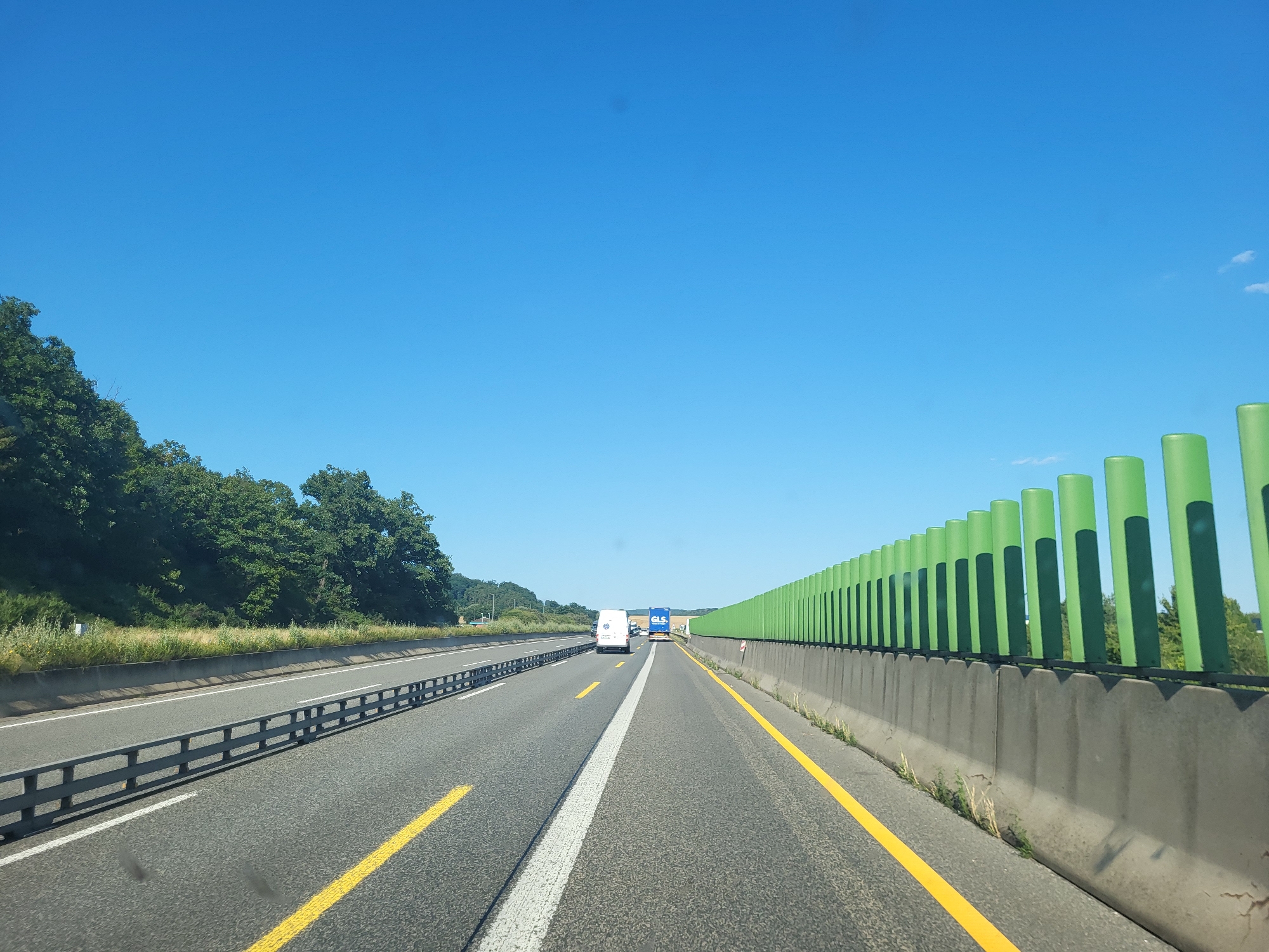 The image depicts a view from a vehicle driving on a highway on a clear, sunny day. The sky is a vibrant blue, suggesting it is daytime, likely late morning or afternoon.
The highway is asphalt with distinct yellow and white lane markings. On the left side of the road, a metal guardrail separates the lanes from a grassy embankment and a dense line of green trees. On the right side, a concrete barrier topped with a row of bright green, cylindrical noise reduction elements creates a striking visual contrast against the blue sky.
Several vehicles are visible on the highway. In the distance, a white van and a blue truck with "GLS" branding are traveling in the same direction as the viewer. The presence of these vehicles indicates that this is an active roadway, likely a major route.
The scene appears to be in a rural or semi-rural area, given the presence of trees and open skies. While the exact location is specified as Laasdorf, Germany, no specific landmarks are clearly identifiable from the image itself, other than the general highway infrastructure. The overall mood is serene and open, conveying a sense of travel and a pleasant day for driving. There is no visible text other than the "GLS" logo on the truck.