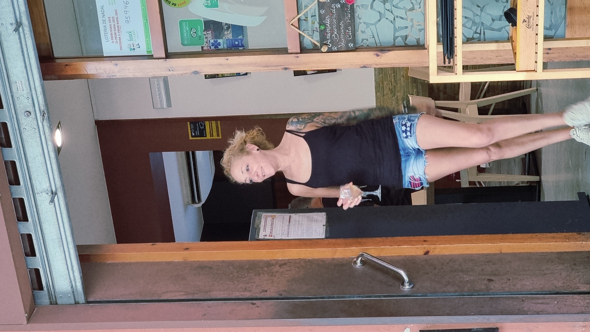 A Caucasian woman with blonde, curly hair is standing in the doorway of a building, holding a stemmed glass of white liquid. She is wearing a black tank top and distressed denim shorts with American flag patches. Her left arm is covered in tattoos. To her right, a wooden shelf unit with various items is visible, including a box labeled "Cacaolat." Above the shelf, a sign with handwritten text and paw prints reads "Animales bienvenidos Humanos ... Cita previa." The environment appears to be an establishment with a rolling metal security gate overhead. The scene is situated in Olot, Spain.