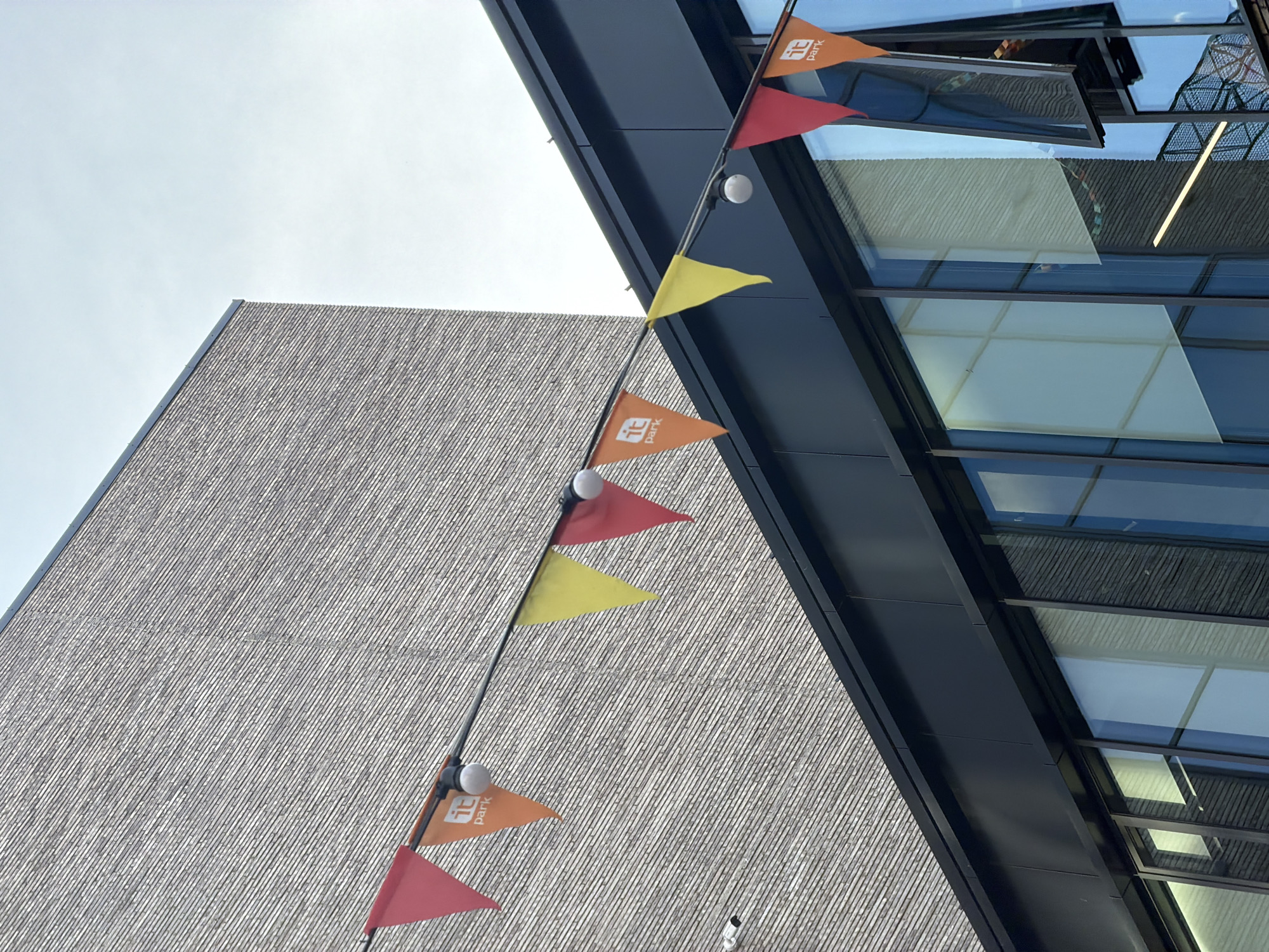 The photograph depicts a low-angle view of building exteriors in Kazan, Russia. The left side of the image shows a building with a light gray, textured facade made up of repeating, narrow, horizontal elements. A dark-colored building stands to the right, featuring a facade of large windows with dark frames. A string of colorful triangular flags, alternately orange (with the word "Park" or "it Park" printed in white), red, and yellow, is strung diagonally across the scene. White bulbs are interspersed along the string of flags. The sky above is overcast. The composition of the image suggests the photograph was taken outdoors, possibly during the day.