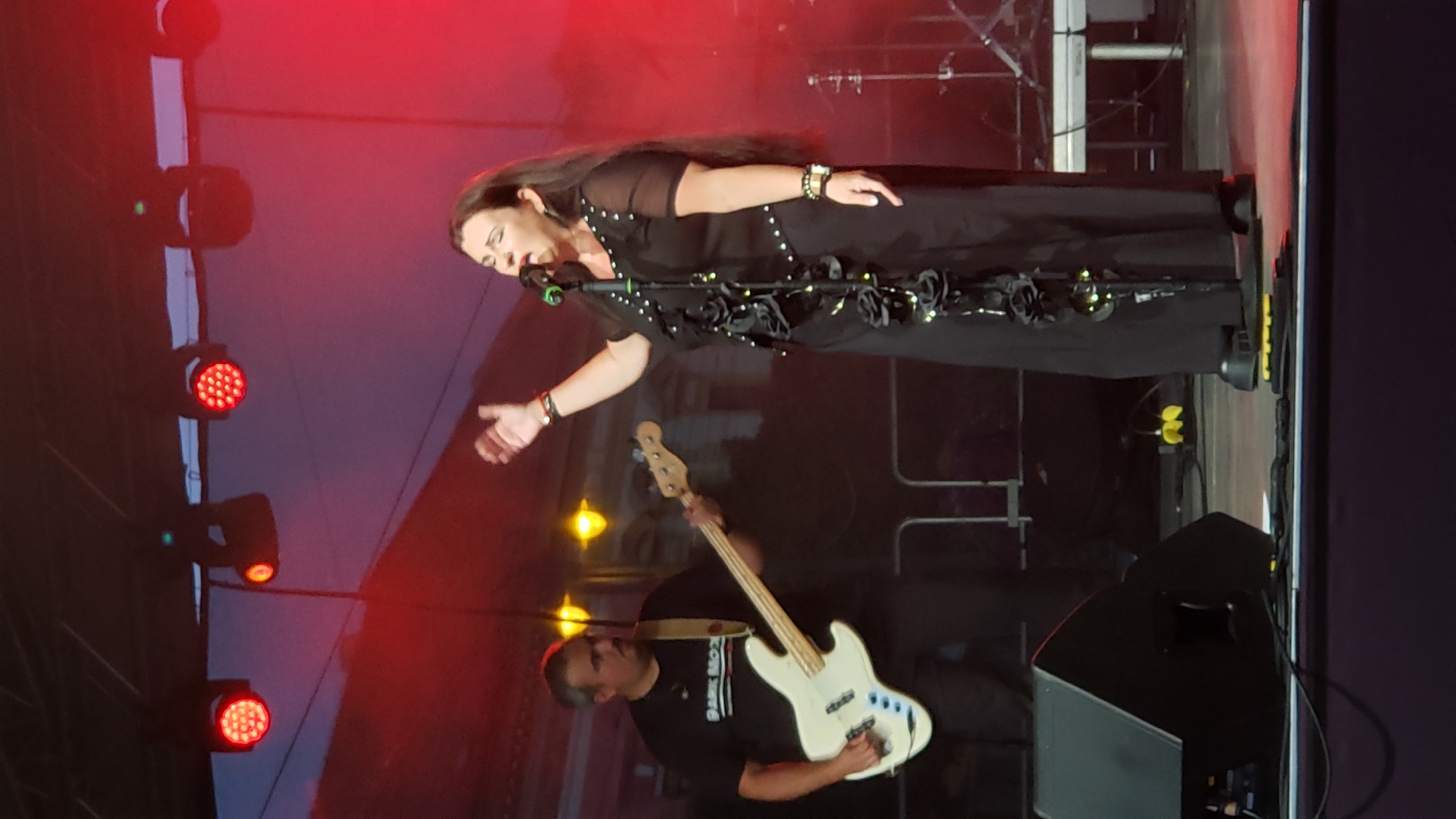 An outdoor concert stage in Prešov, Slovakia, is illuminated by red stage lights. On the right, a woman with long dark hair is singing into a microphone, wearing a long black dress adorned with black roses and studs. She has a bracelet on her right wrist. To her left, a man with dark hair and a mustache is playing a white electric bass guitar. He is wearing a black t-shirt with white text that reads "DARK MOO". In the foreground, a large black speaker is visible, and yellow stage equipment is scattered on the stage floor. The background is dark with hints of buildings and streetlights, suggesting an urban setting.