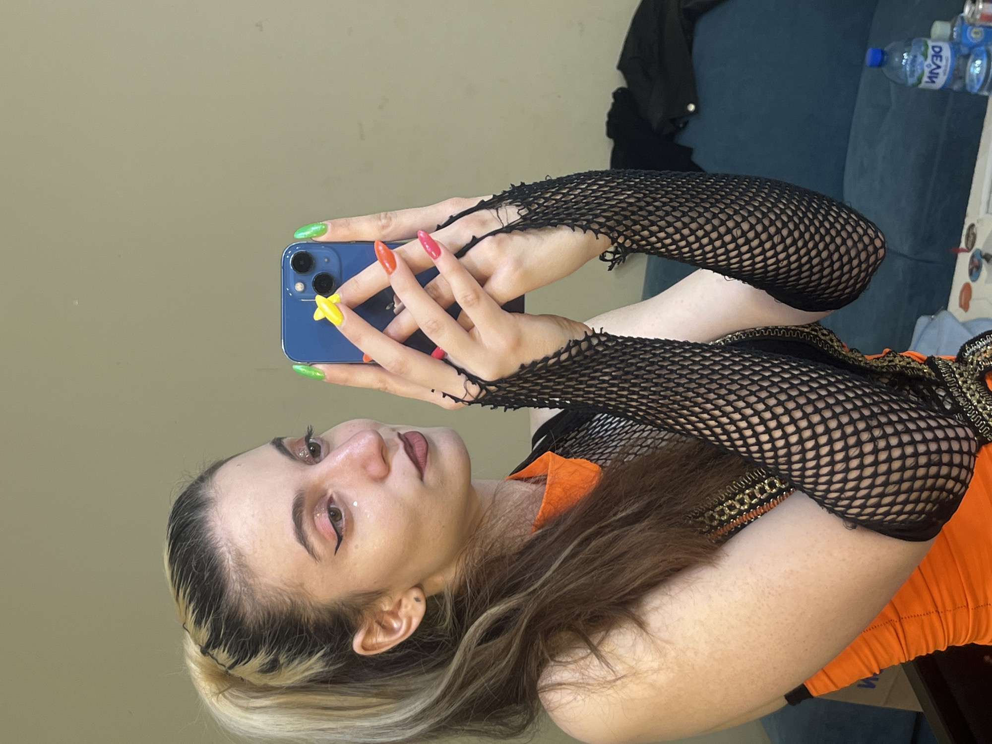 A person is taking a selfie with a blue phone. The person is wearing black fishnet gloves and an orange top. The person's nails are painted in different colors. The background is a beige wall and a blue sofa. There are bottles of water on the sofa. The location is not discernible as Plovdiv, Bulgaria from the image.