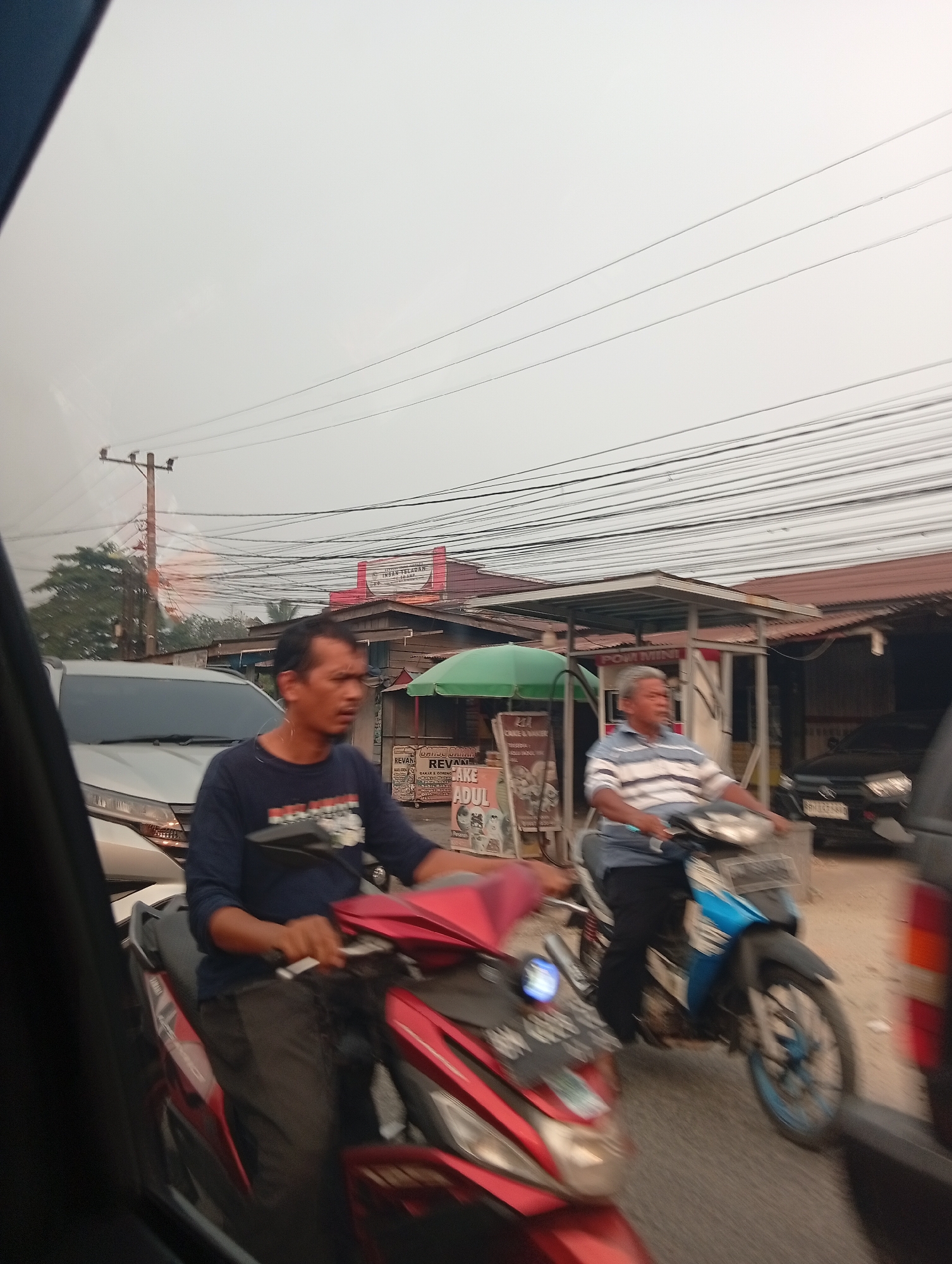 The image depicts a street scene in Tarai Bangun, Indonesia, with a considerable amount of overhead power lines. Two men on motorcycles are visible in traffic. The man in the foreground, wearing a dark blue shirt, is on a red and black motorcycle. The man behind him, wearing a striped blue and white shirt, is on a blue and black motorcycle. Several vehicles, including cars and other motorcycles, are present in the background, suggesting moderate traffic congestion. Commercial buildings with red roofs line the street, and a green umbrella is visible near one of the buildings. The sky is overcast, indicating potentially hazy or cloudy weather. The overall scene captures a moment of daily life in a bustling Indonesian town.
