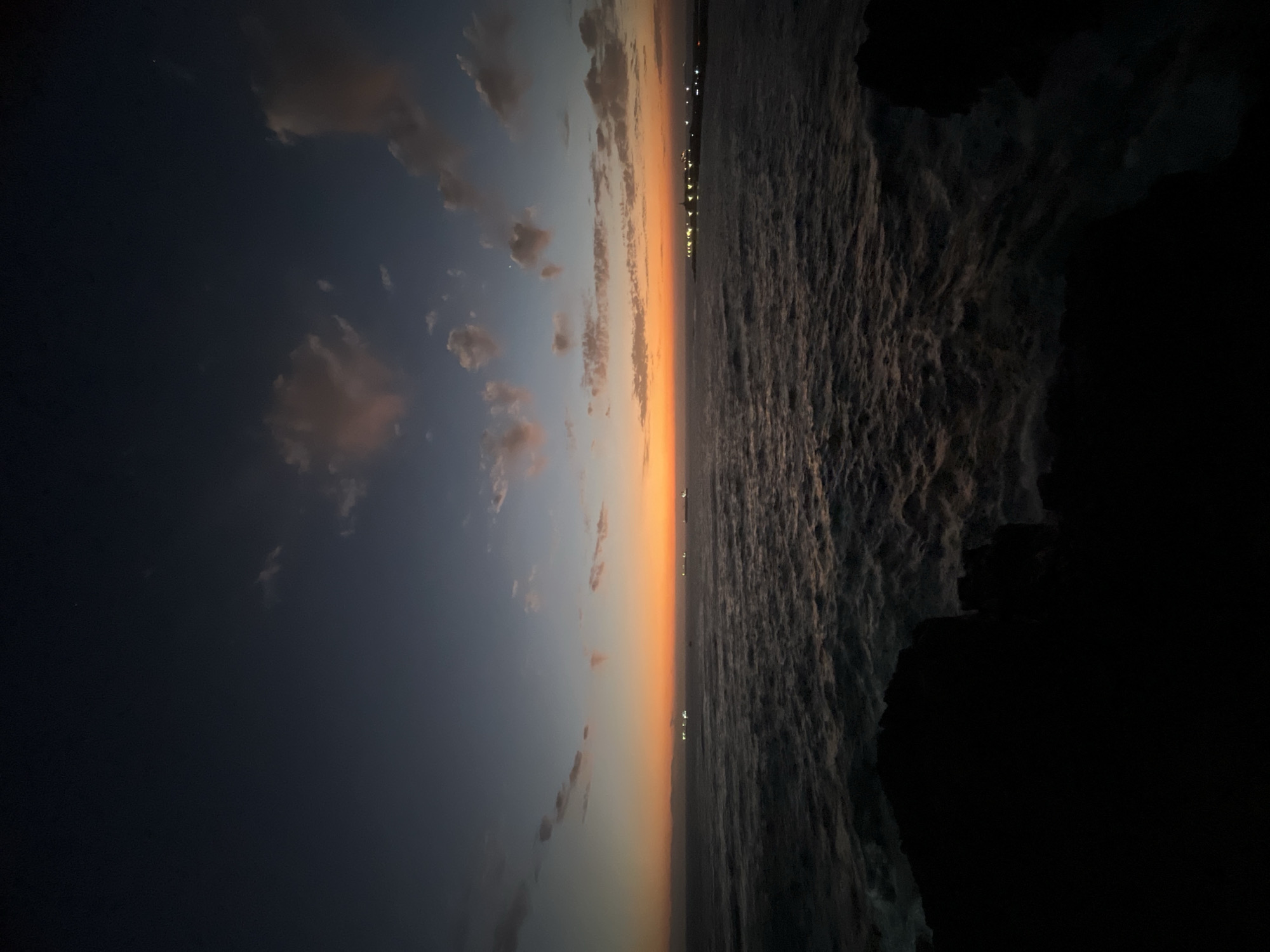The photograph captures a sunset scene in Arica, Chile. The sky transitions from dark blue on the left to a bright orange gradient near the horizon. Wispy clouds are scattered across the sky. Lights, likely from the city, are visible along the horizon. The ocean, textured by waves, occupies the right side of the frame. The shoreline is visible on the right, fading into darkness.