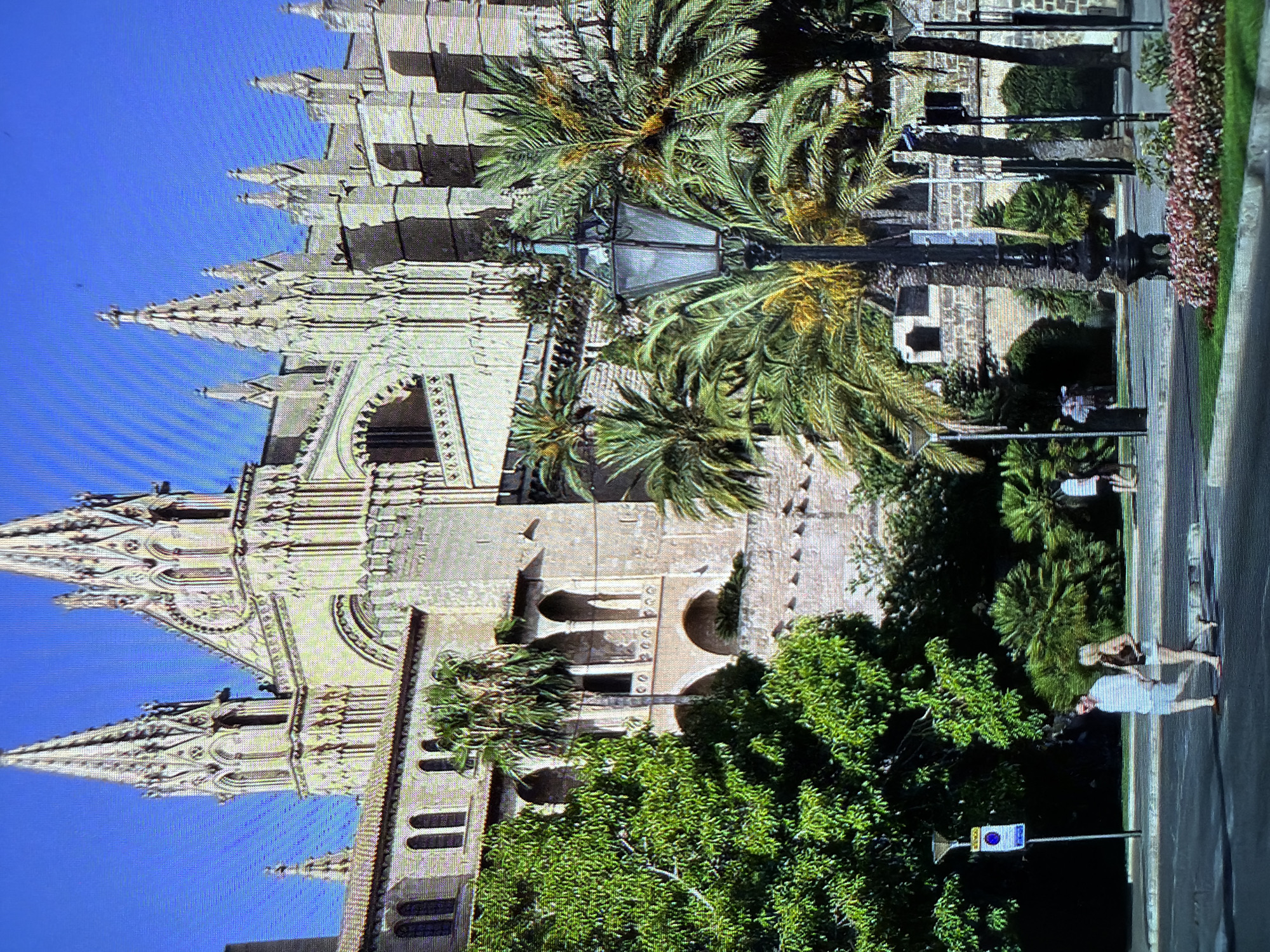 This media file captures a sun-drenched scene featuring a large, ornate Gothic-style building with spires and intricate architectural details, likely a cathedral or similar historical structure. Lush green trees and palm trees with vibrant foliage surround the building, creating a striking contrast with the stone architecture.
Several people are visible in the foreground, walking along a paved path or street adjacent to the building. One person is wearing a light-colored top and dark shorts, appearing to be walking away from the camera. Another individual, seen from the side, is wearing a light-colored top and lighter shorts. A third person is visible further down the path.
A streetlamp with a dark metal frame and glass panes stands prominently amidst the palm trees. A blue and white sign with circular symbols is mounted on a pole near the people.
The bright sunlight and clear blue sky suggest it is daytime with fair weather. The presence of palm trees and the architectural style hint at a Mediterranean or similar warm climate, aligning with the provided location context of Sant Antoni De Portmany, Spain. The overall atmosphere appears relaxed and leisurely, with people enjoying the outdoor environment.