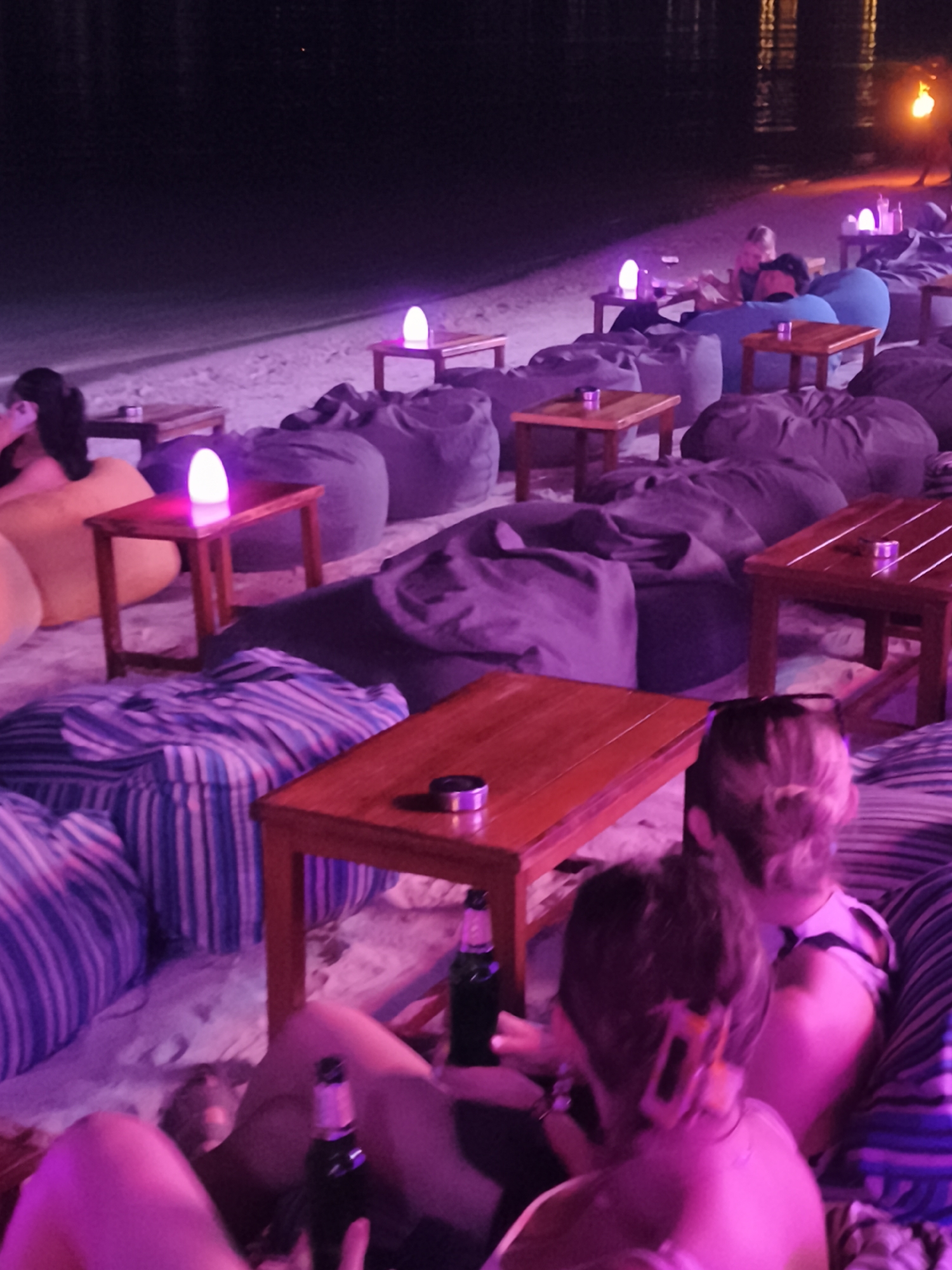 At night on a beach in the Ko Samui District, Thailand, several people are seated on beanbag chairs and cushions around small wooden tables. The scene is illuminated by purple and pink ambient lighting, including small lamps on the tables. Several bottles, possibly of beer, are visible on the tables and in people's hands. The atmosphere appears relaxed and social, suggesting a bar or lounge setting on the beach. The water is visible in the background, reflecting the lights.