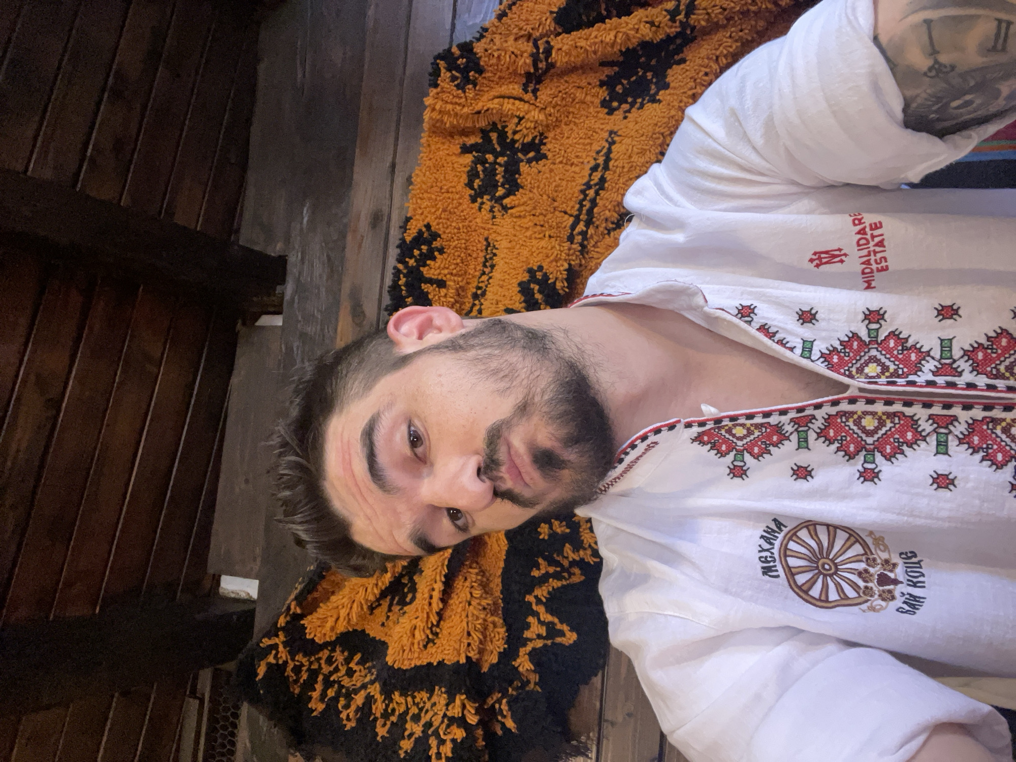 A low-angle, medium shot captures a man with dark hair, a beard, and a mustache posing for a selfie. He is looking directly at the camera with a slight pout and his head is tilted to the left. He is wearing a white shirt with red and black embroidered detailing around the collar and down the front. The shirt has a circular logo on the left chest with the word "Mekhana" above a depiction of a wagon wheel and some floral elements, with "Bay Koce" written below. On the right chest, there is a logo with the letters "M" inside a shield and the words "Midalidare Estate" below.
The man is positioned in front of a large, fuzzy, orange and black textile with a tree-like pattern. Behind him and above, wooden beams and planks form a rustic ceiling. The background appears to be an indoor setting with wooden walls. The lighting is natural and appears to be coming from the front, illuminating the man's face. There is a visible tattoo on the man's right arm.
