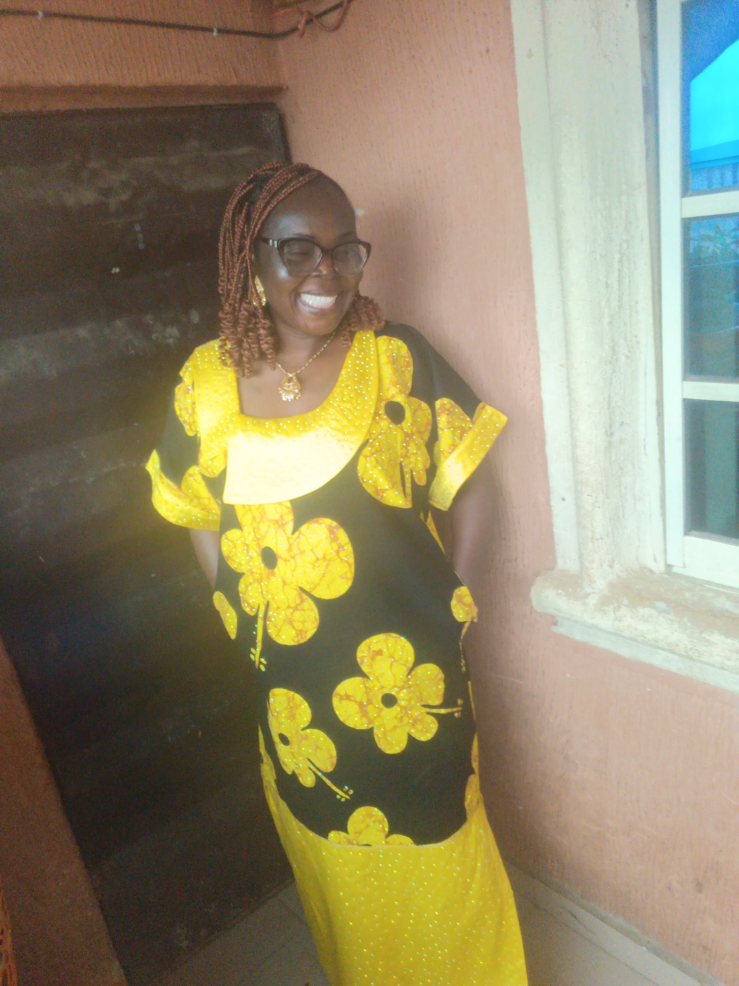 A Black woman stands in front of a textured pink wall and a window. She is wearing a yellow and black dress with a floral pattern and a necklace. Her eyes are closed, and she is smiling. She has braided hair. The image appears to be taken indoors, possibly in a residential area in Onitsha, Nigeria.