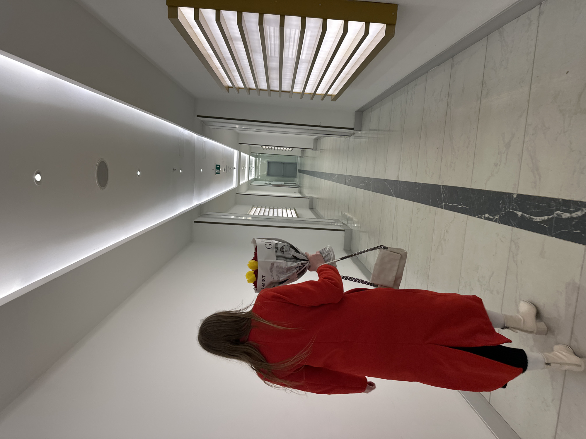 A woman wearing a red coat and white boots walks down a long, brightly lit hallway, holding a bouquet of flowers wrapped in newspaper. The hallway has white walls, a white tiled floor with a dark marble-like accent strip, and a modern linear light fixture overhead. The location is a brightly lit hallway in a building in the city of Batumi, Georgia.