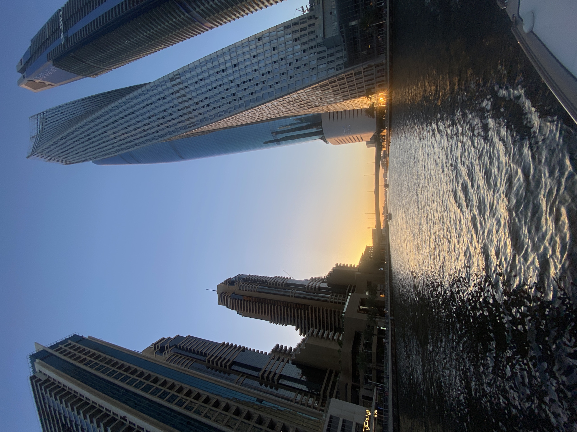The image captures a scene in Dubai, United Arab Emirates, featuring a combination of modern architecture and water. Skyscrapers dominate the frame, some appearing to lean into the shot, their glass facades reflecting the sky and the setting sun. The architectural style includes tall, rectangular buildings with repeating window patterns and stepped designs. Below, water reflects the golden light of the sunset, creating rippling patterns and highlights. The buildings along the water's edge include both low-rise structures and taller ones. The sky transitions from blue to a gradient of orange and yellow near the horizon, indicating either sunrise or sunset. Overall, the scene portrays the modern urban landscape of Dubai set against the backdrop of a water body and a vibrant sky.