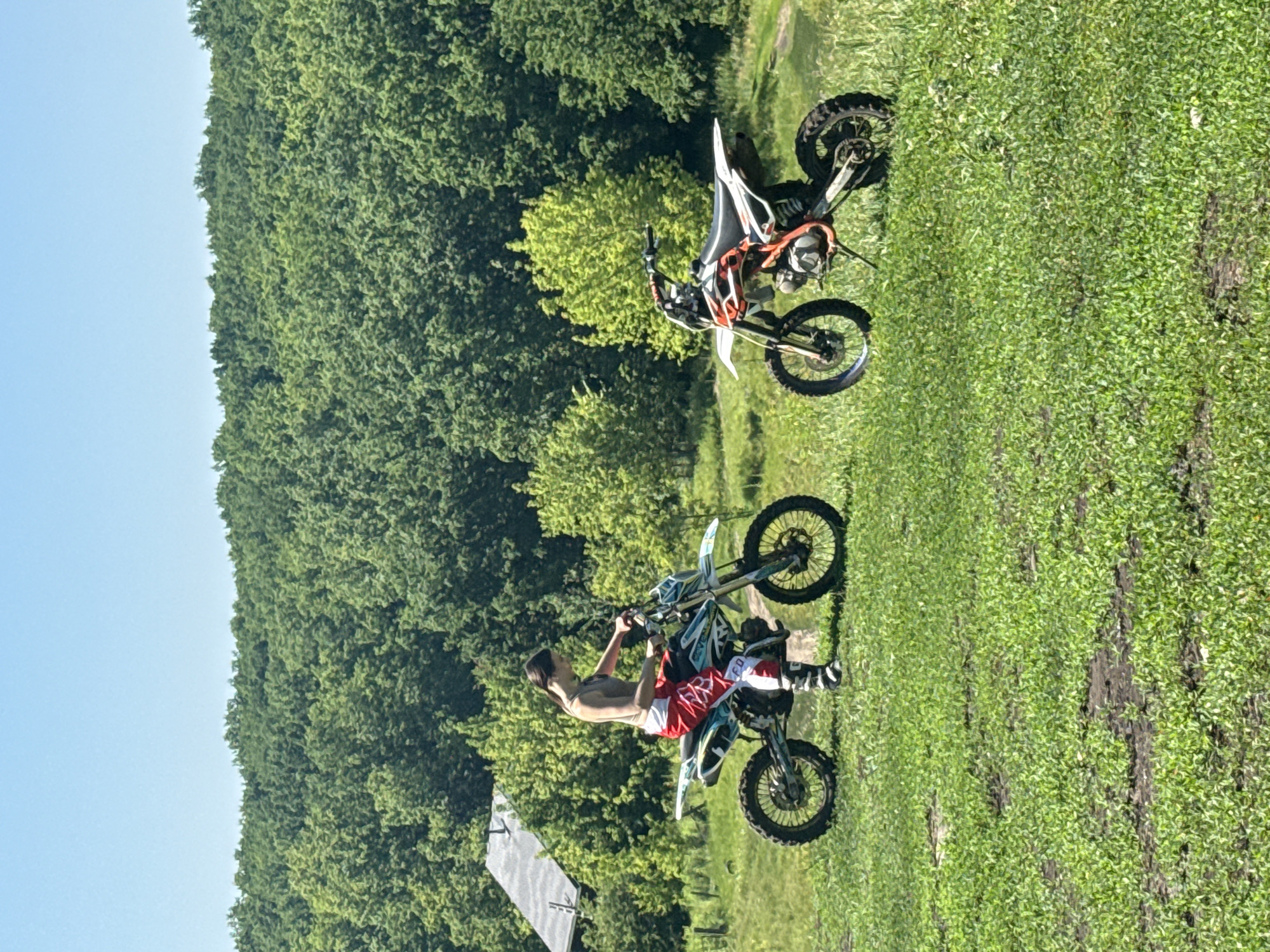 In Novokuybyshevsk, Russia, two individuals are engaged in motocross activities on a sunny day. The scene is set in a grassy field with a lush green forest in the background. The sky is clear and blue, indicating fair weather.
In the foreground, a person is performing a stunt on a dirt bike. They are wearing a tank top and red and white motocross pants. The dirt bike is predominantly white with blue and black accents and has "FOX" visible on the pants. The rider is in mid-air, suggesting an acrobatic maneuver.
Behind the first rider, another dirt bike is parked. This bike is white with orange and black detailing. It appears to be a different model than the one being ridden in the foreground.
The environment suggests an outdoor recreational area, possibly a motocross track or a designated riding spot, with natural terrain. The overall impression is one of action and outdoor sport. There are no other notable emotions or details conveyed besides the activity itself. The time of day appears to be daytime, likely late morning or afternoon, given the bright sunlight and clear sky.