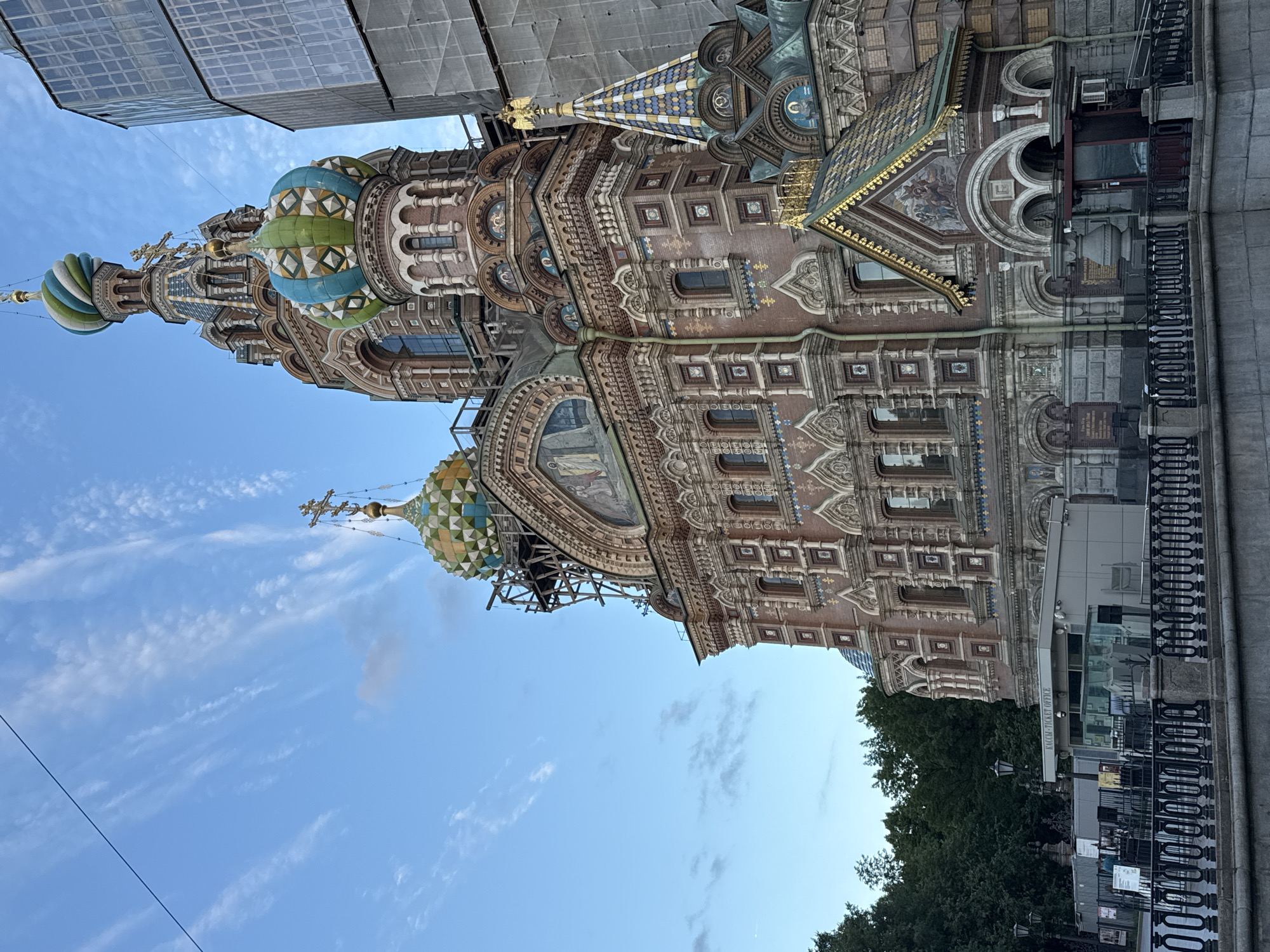 The image shows the Church of the Savior on Spilled Blood in Saint Petersburg, Russia. The ornate church is characterized by its colorful onion domes and detailed brickwork. In the foreground, a black metal fence separates the sidewalk from a canal. Behind the fence, there is a ticket office with glass windows and signs. The church itself is a dominant feature, with scaffolding visible on some parts of its exterior, suggesting ongoing restoration or construction work. To the right of the church, a modern building with a glass facade is under construction, covered in scaffolding and grey sheeting. The sky above is a vibrant blue with wispy white clouds. Trees are visible in the background on the left side of the image.