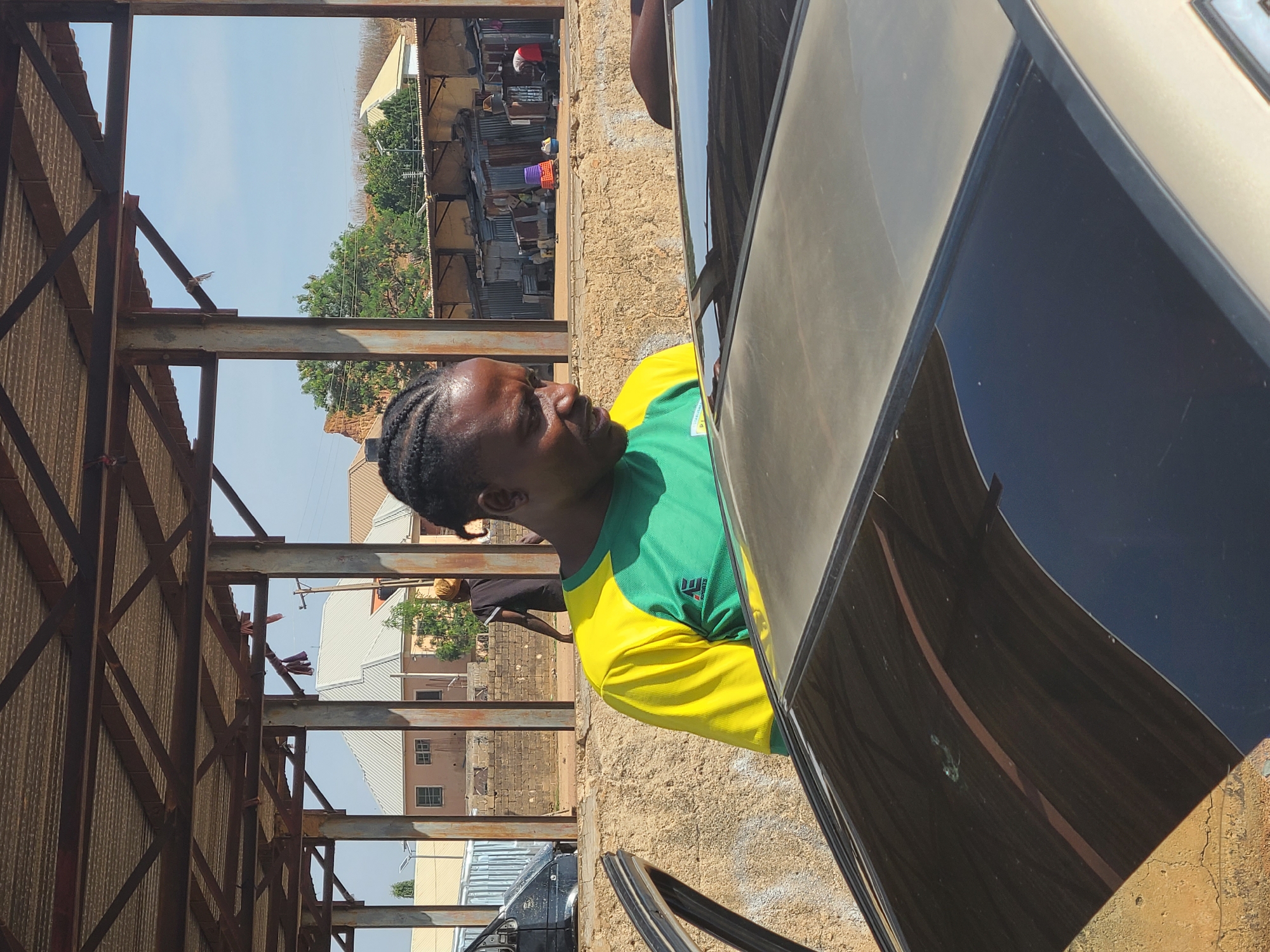 A man with braided hair, wearing a green and yellow shirt, stands beside a shiny, dark-colored vehicle. The scene is in Jos, Nigeria, suggested by the architectural style and the presence of corrugated metal structures. The man is positioned outside a metal frame building, possibly under construction or a market stall. In the background, a residential area with trees and a cluster of buildings is visible. Some people are also visible in the background near the buildings. The ground is dusty and uneven. The shiny surface of the vehicle reflects the surroundings.