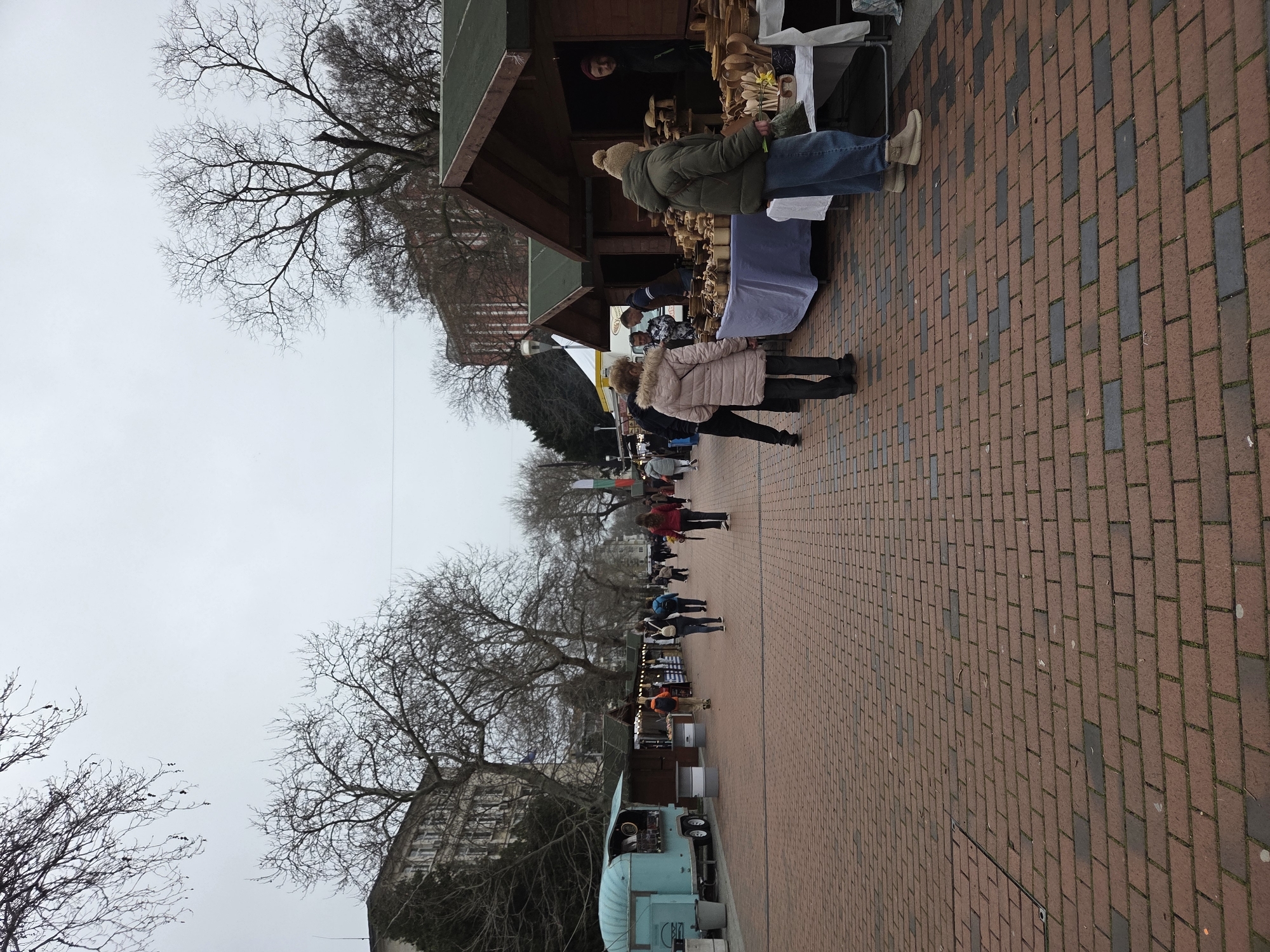 The image depicts a bustling street scene in Varna, Bulgaria, likely a market or fair. The ground is paved with red and dark bricks, creating a distinct pattern. Several market stalls line the street, displaying various goods, including wooden items. People of different ages and attire are browsing and interacting with vendors. There are leafless trees on either side of the street. A vintage, light blue truck/van is parked near the stalls. In the background, there is a building and the sky appears overcast. A Bulgarian flag is seen further down the street.