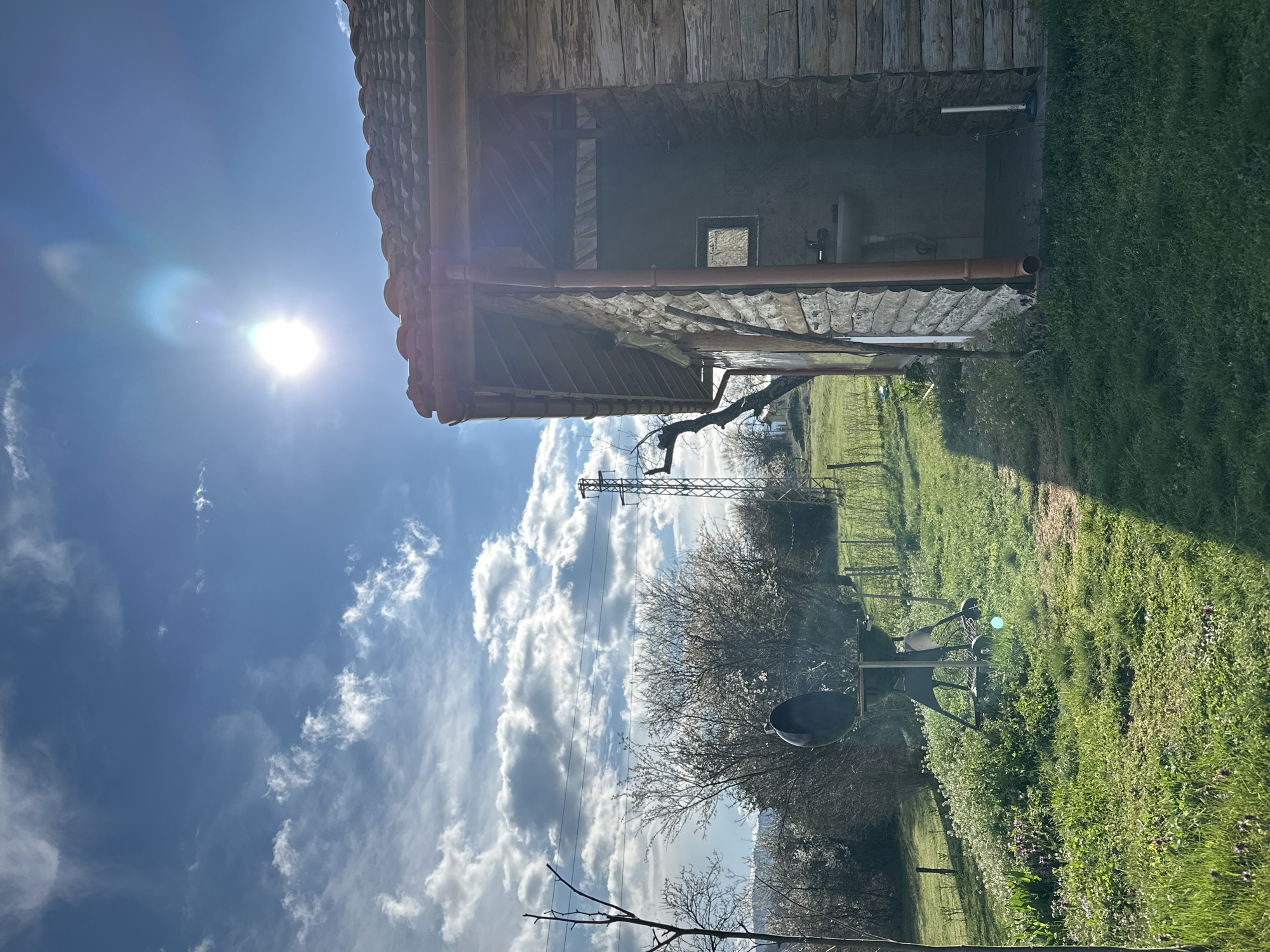 The image shows a rural scene near Mrachenik, Bulgaria. A small, rustic building with a tiled roof and wooden walls stands on the edge of a grassy field. A metal utility pole is visible in the background, behind a tree with white blossoms. In the foreground, a charcoal grill sits in the grass. The sky is blue with scattered white clouds, and the sun is shining brightly.