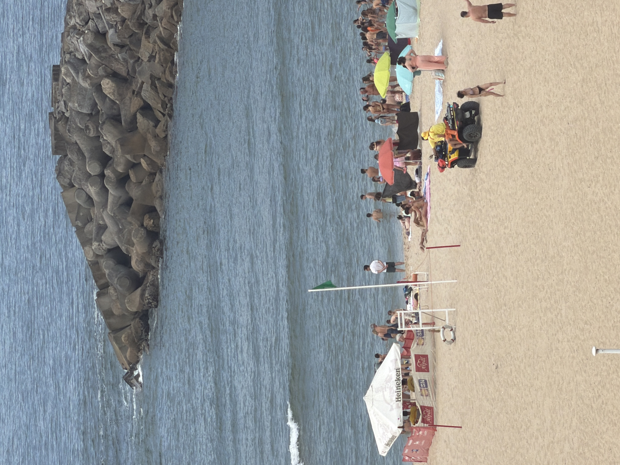This is an aerial, medium-shot photograph of a crowded beach in Espinho, Portugal, on a hazy day. The scene is dominated by the sandy beach in the foreground, the choppy blue ocean in the midground, and a large, concrete breakwater made of tetrapods in the background. Numerous beachgoers are spread across the sand, many in swimwear, engaging in various activities. Some are lying on towels, others are standing and talking, and a group is partially submerged in the water. Beach umbrellas, a white tent with "Heineken" branding, and a red safety barrier are visible on the sand. A green flag is planted on a pole near the water's edge. In the mid-right of the frame, a yellow ATV with two individuals in yellow vests, likely lifeguards, is parked on the sand. Several beach umbrellas in red, yellow, and green hues are scattered amongst the beachgoers. The breakwater, constructed of numerous interlocking concrete tetrapods, extends into the ocean, protecting the coastline. The overall impression is of a busy summer day at the beach.