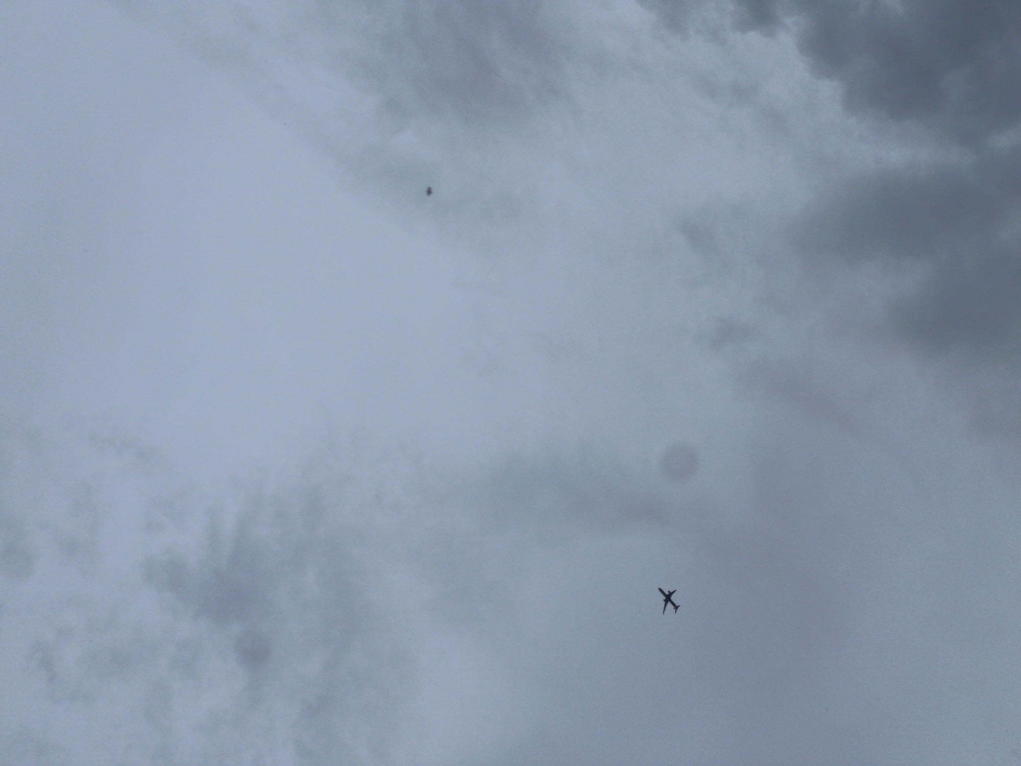 A gray sky with scattered clouds. An airplane is visible in the lower right quadrant and another small object is in the upper middle section.