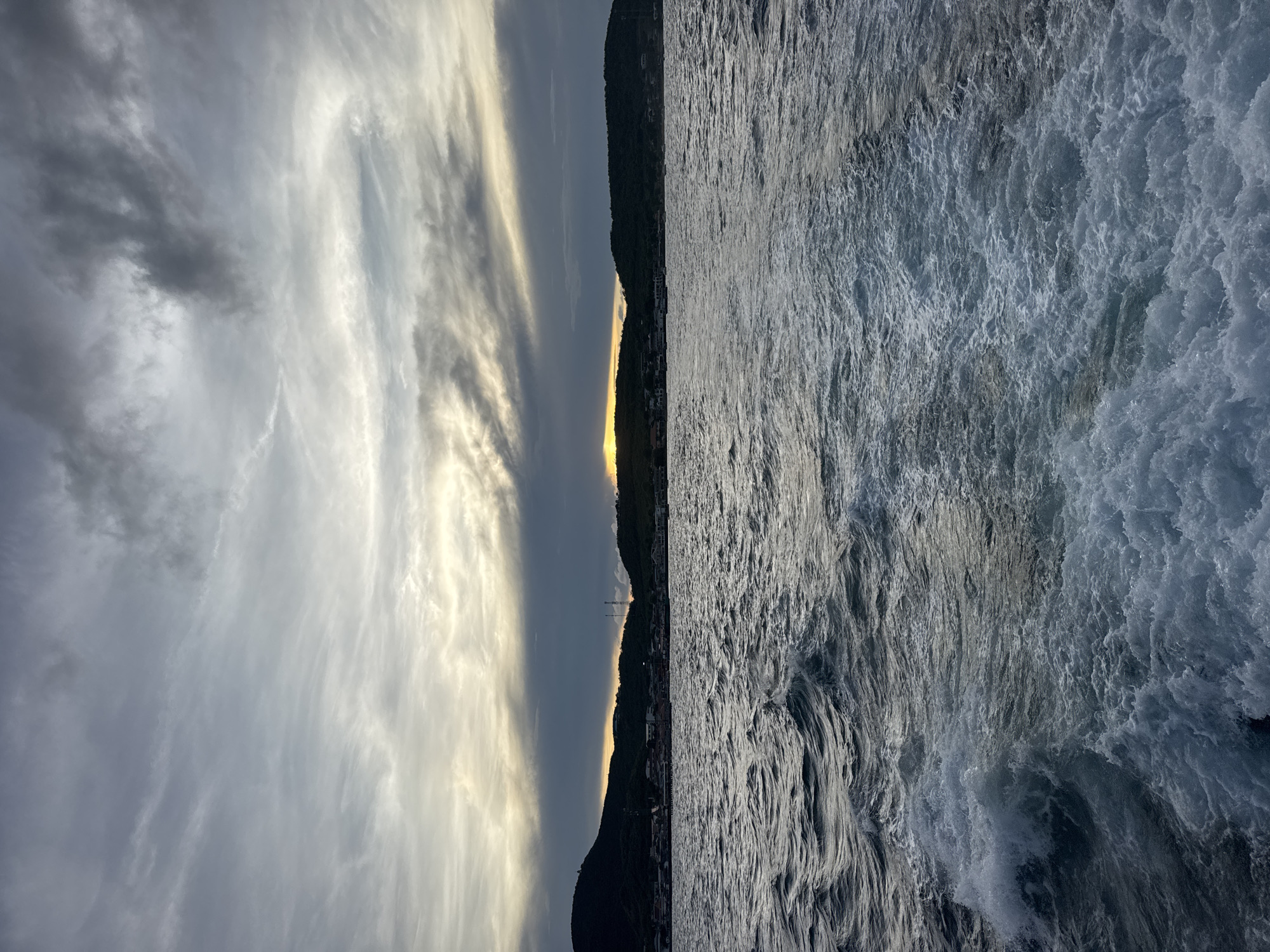 The image shows a seascape with a dramatic sky. On the right, turbulent sea with white foam crests reflecting sunlight. A dark silhouette of a coastline runs horizontally across the center. Between the coastline and the sky, a bright golden horizon hints at sunset or sunrise. The sky is a mix of grey and white clouds. The coastline appears to be a settled area with buildings and possibly a communication tower. The location is Pattaya City, Thailand.