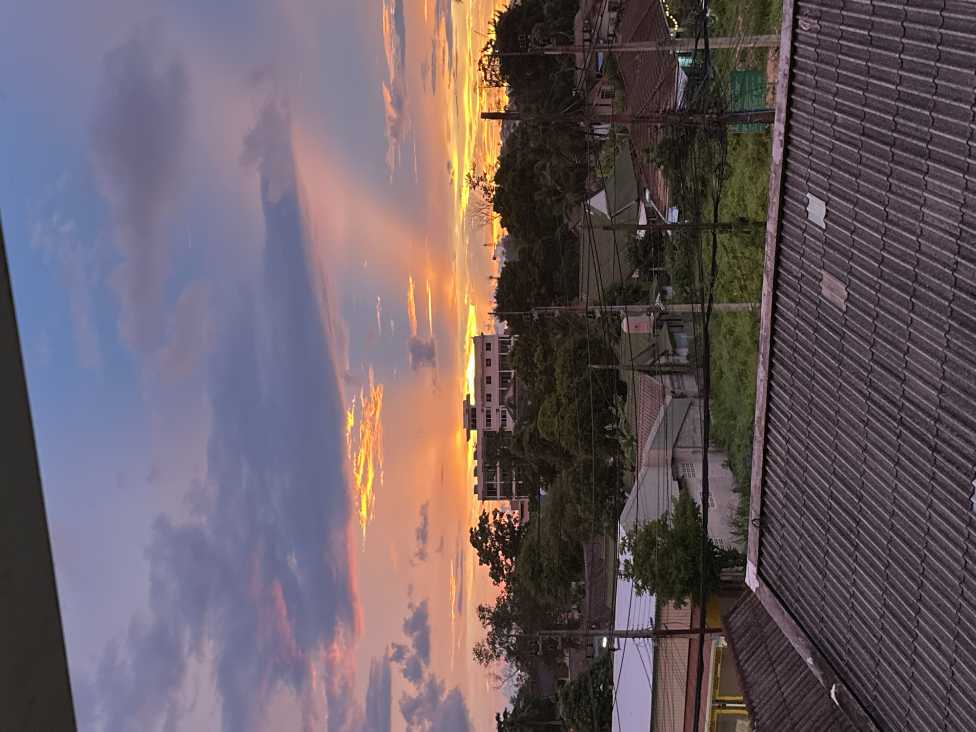 The image depicts a sunset over a residential area in Karon, Thailand. The sky is a vibrant display of orange, pink, and yellow hues, with streaks of light piercing through the clouds. The scene is dominated by the warm glow of the setting sun, casting long shadows over the buildings and trees.
In the foreground, the tiled roof of a house is visible, with its textured surface capturing the fading light. Beyond the roof, a cluster of trees forms a dense green canopy, interspersed with utility poles and electrical wires that crisscross the landscape. Several buildings of varying heights and architectural styles are scattered throughout the area, including a multi-story building with white facades and numerous windows. The overall impression is one of a quiet, everyday scene bathed in the dramatic beauty of a tropical sunset.
The time of day is clearly dusk, as indicated by the low position of the sun and the warm colors in the sky. The weather appears clear, with the clouds playing a crucial role in shaping the visual spectacle of the sunset. While no people are explicitly visible, the presence of houses and utility infrastructure suggests a populated, inhabited area. The image evokes a sense of peace and tranquility, characteristic of a serene evening in a tropical setting. There is no visible text in the image.