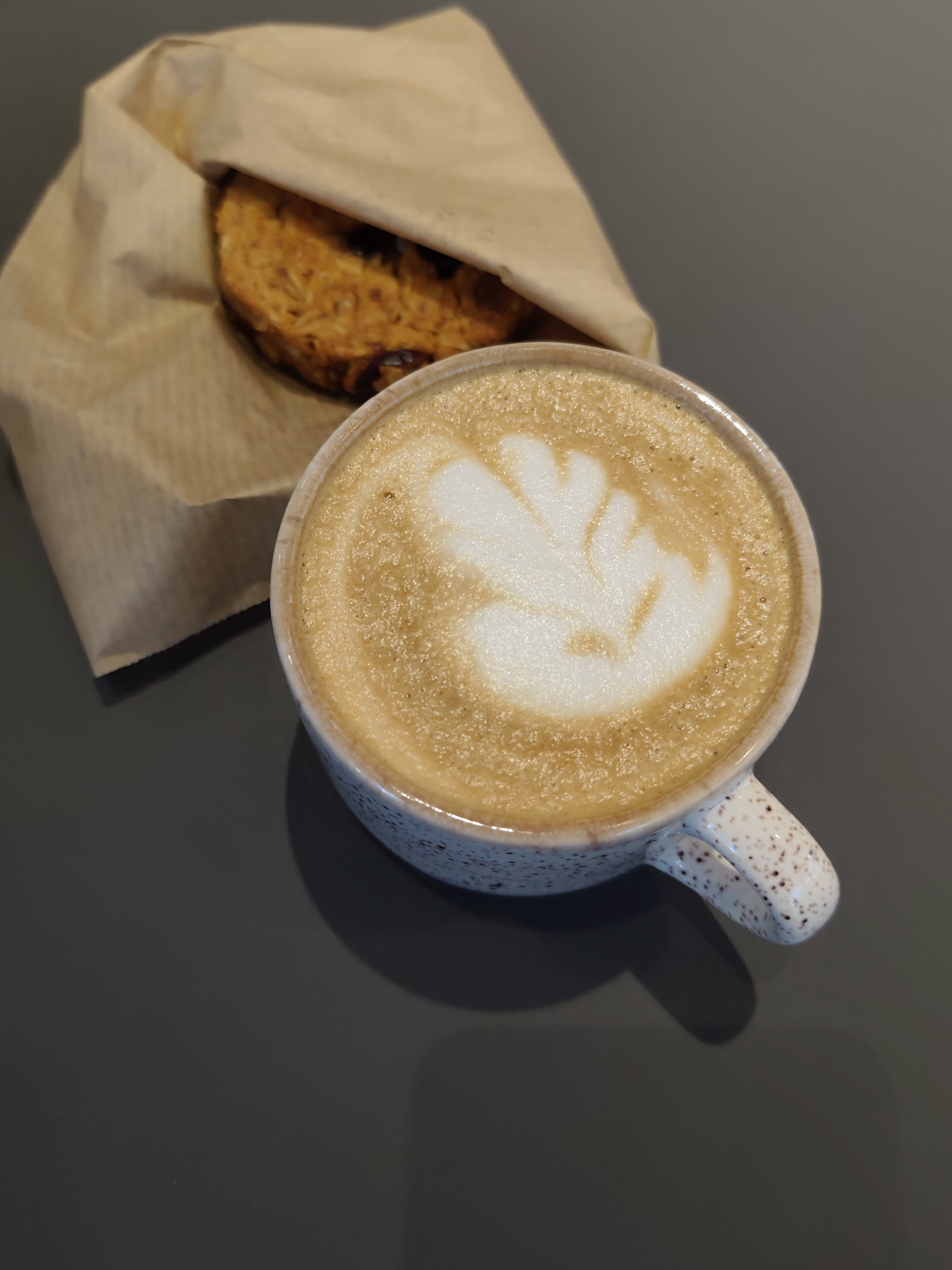 A close-up, high-angle shot shows a cappuccino with latte art and a piece of bread wrapped in brown paper, both resting on a dark gray surface. The coffee cup is white with small brown speckles and a handle. The latte art is a white foam pattern on top of the brown coffee. The bread, partially visible, appears to be a round, flatbread with visible raisins or dried fruit. The lighting is soft and casts a subtle shadow from the coffee cup. The scene is devoid of any people or activity, suggesting a quiet moment of consumption. The location in Sochi, Russia, is not visually indicated by any specific landmarks or cultural cues within the frame.