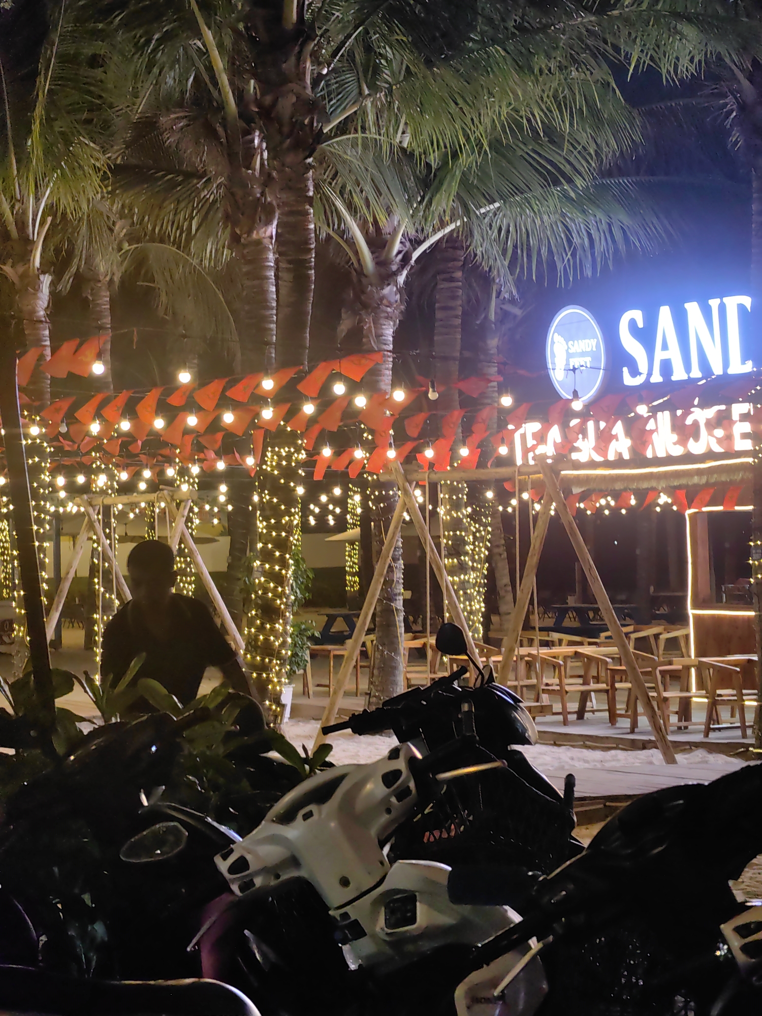The image shows an outdoor scene at night, likely in Bai Chay Ward, Vietnam, given the presence of "Sandy Foot Tequila House" signage. The foreground is dominated by parked motorbikes, with a person visible behind some foliage. The midground features a bar or restaurant area decorated with string lights and red pennants. Tables and chairs are visible, suggesting a seating area. Palm trees are present in the background, adding to the outdoor ambiance. The bar has a neon sign that displays the name "Sandy Foot Tequila House". The scene is generally illuminated by the decorative lighting and the bar's signage.