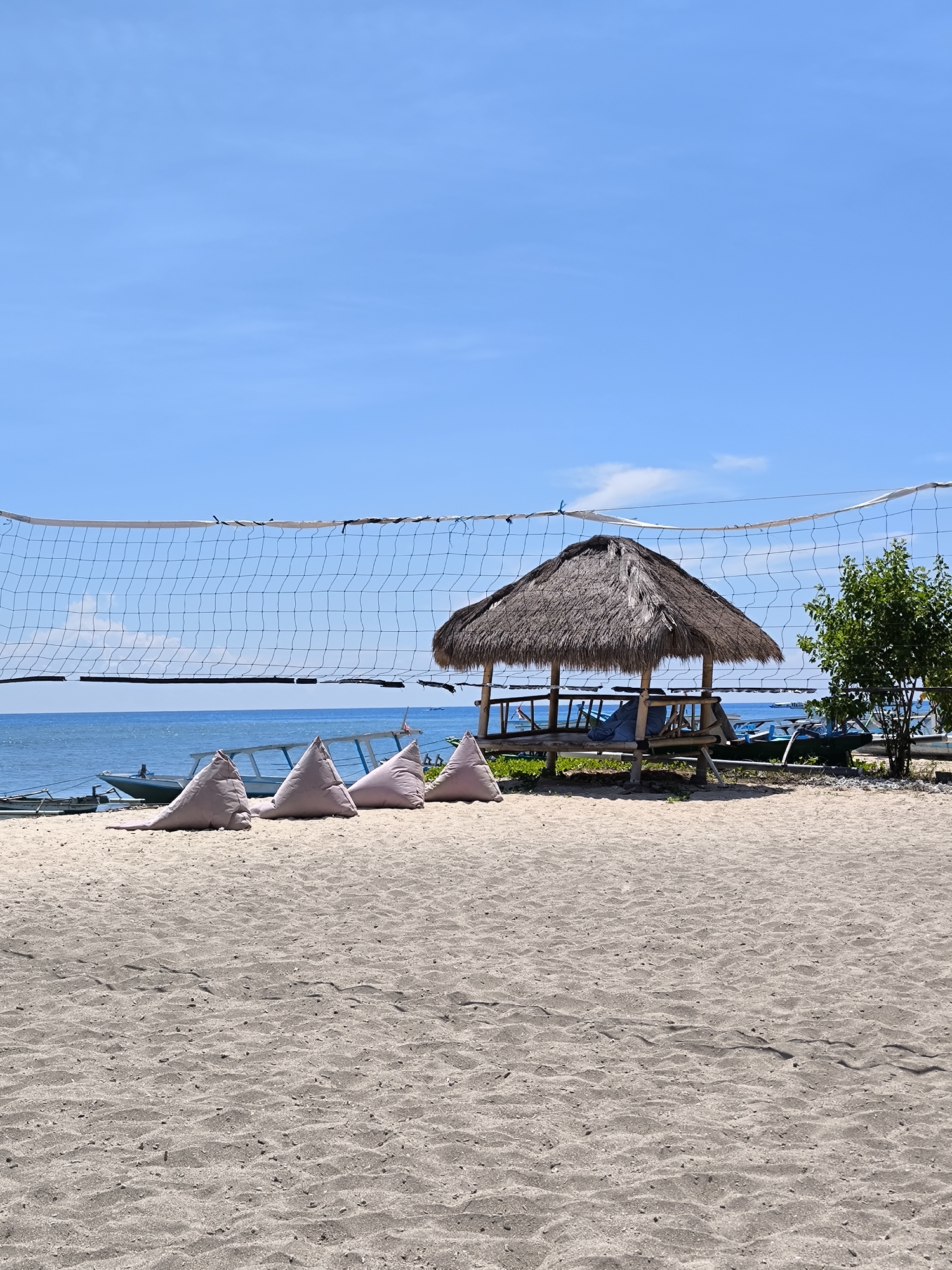 The image depicts a beach scene in Unknown, Indonesia. The foreground features a sandy beach with textured patterns. A volleyball net stretches across the frame. Behind the net are three triangular cushions placed on the sand. A small hut with a thatched roof is situated to the right, providing shade. A small tree grows next to the hut. In the background, a calm blue sea is visible, with several boats anchored. The sky is clear and blue.
