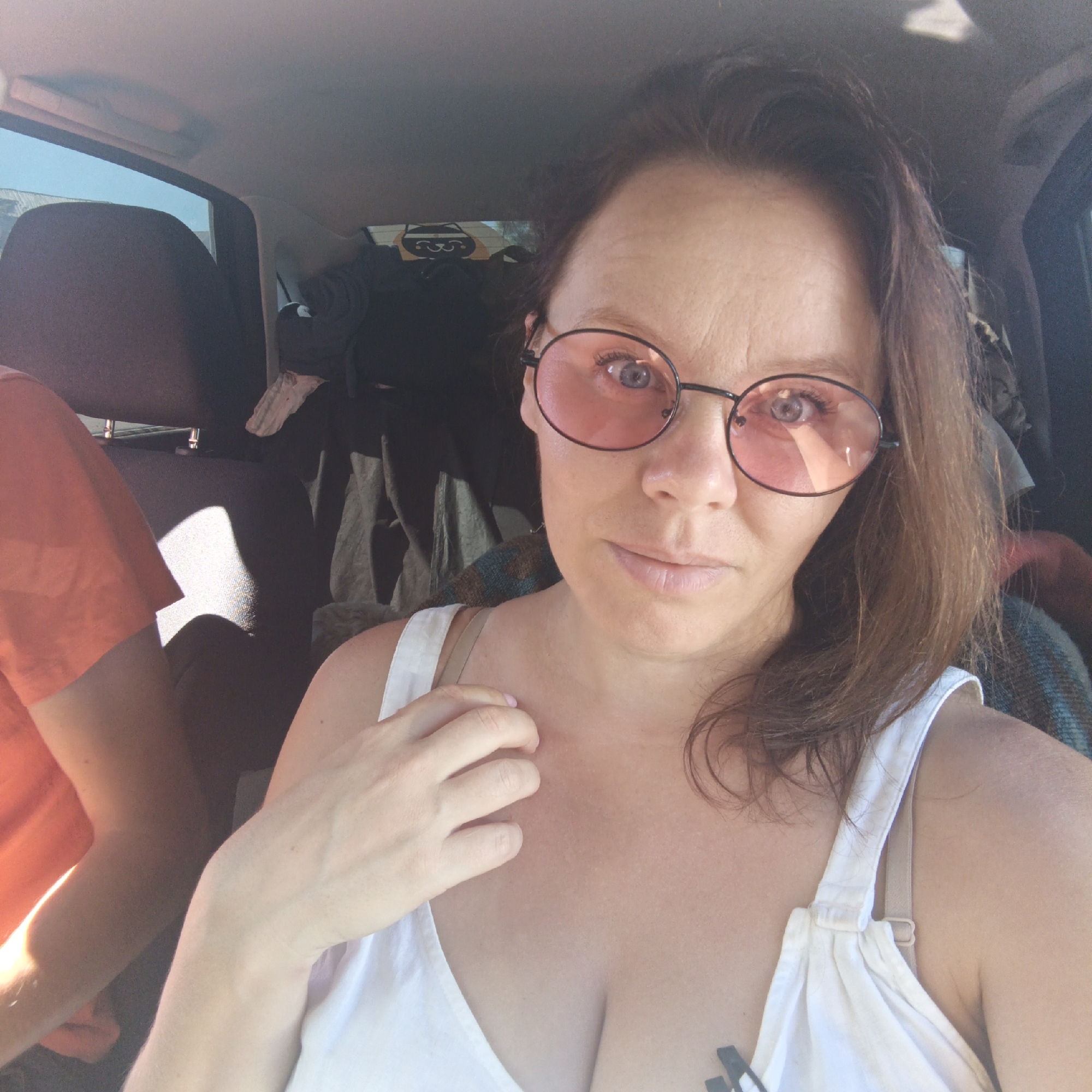 The image is a selfie taken from inside a vehicle, presumably a car, on a sunny day. The main subject is a woman with brown hair and blue eyes, wearing round pink-tinted glasses and a white tank top. She is looking towards the camera with a slight smile. Her right hand is resting on her chest, near her shoulder. To the left of the woman, another person is visible from the shoulders up, wearing an orange t-shirt. The interior of the car is filled with various items, including what appears to be luggage or bags, suggesting a journey or travel. The lighting suggests it is daytime, with sunlight coming in through the windows, casting shadows. The background shows the interior of the car, including headrests and the ceiling. There is a sticker with a cartoonish design on the rear window. The location is identified as Tatarskii Saraktash, Russia, though this information cannot be verified from the visual content alone.