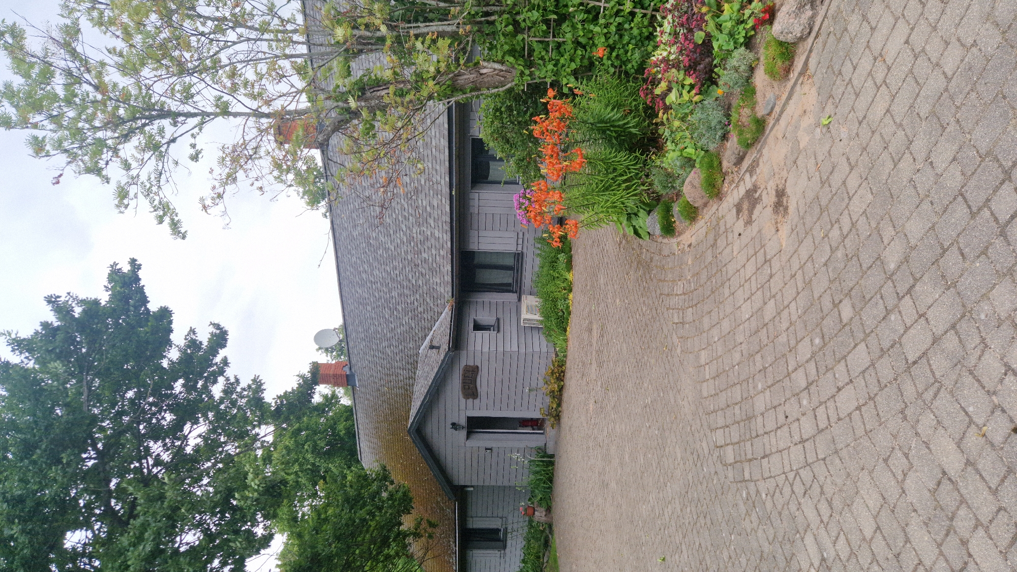 The image depicts a grey, single-story house with a grey shingle roof, set within a natural landscape. A paved driveway made of grey interlocking bricks curves towards the entrance of the house. The house features multiple windows and a prominent wooden sign above the entrance reading "LERUJI". To the right of the entrance, an air conditioning unit is visible. The area surrounding the house is landscaped with a variety of plants and flowers, including bright orange lilies and purple blooms, along with large rocks. Mature trees with green foliage are present on the left and right sides of the house. The sky is overcast with a few visible chimneys and a satellite dish atop the roof. This scene is located in Vārzas, Latvia.