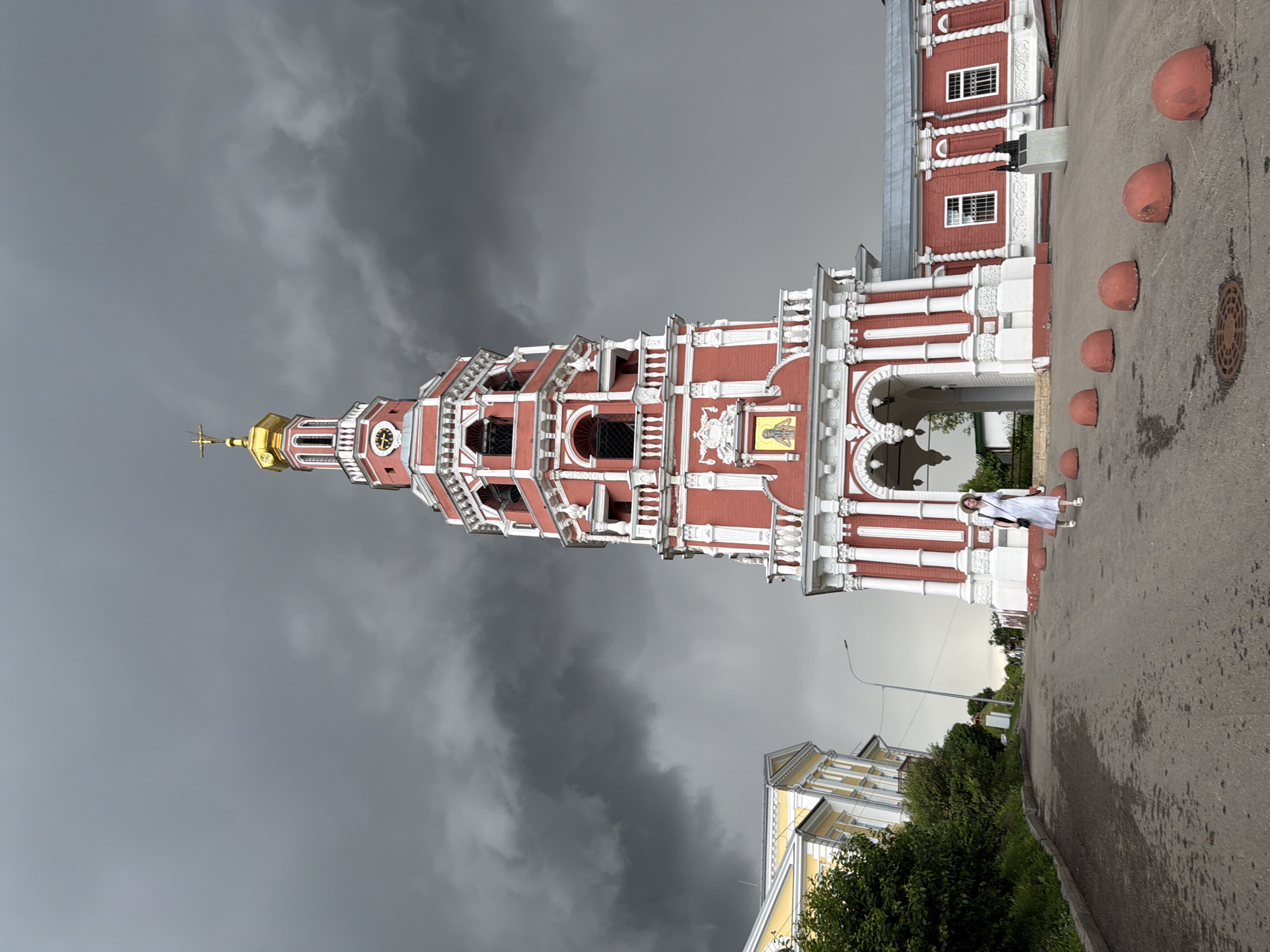A woman in a light-colored dress and hat stands on an asphalt path next to a red and white bell tower. The bell tower has intricate architectural details, including arches, columns, and decorative elements. A golden cross tops the tower, and a clock face is visible. The sky above is dark and stormy, suggesting overcast weather. In the background, another building with yellow and white detailing is partially visible, along with lush green trees. Several red, dome-shaped bollards line the path near the bell tower. The scene appears to be outdoors, possibly in a historical or religious complex in Nizhny Novgorod, Russia. The lighting and dark clouds evoke a sense of impending rain or a dramatic atmosphere. No visible text is present in the image.