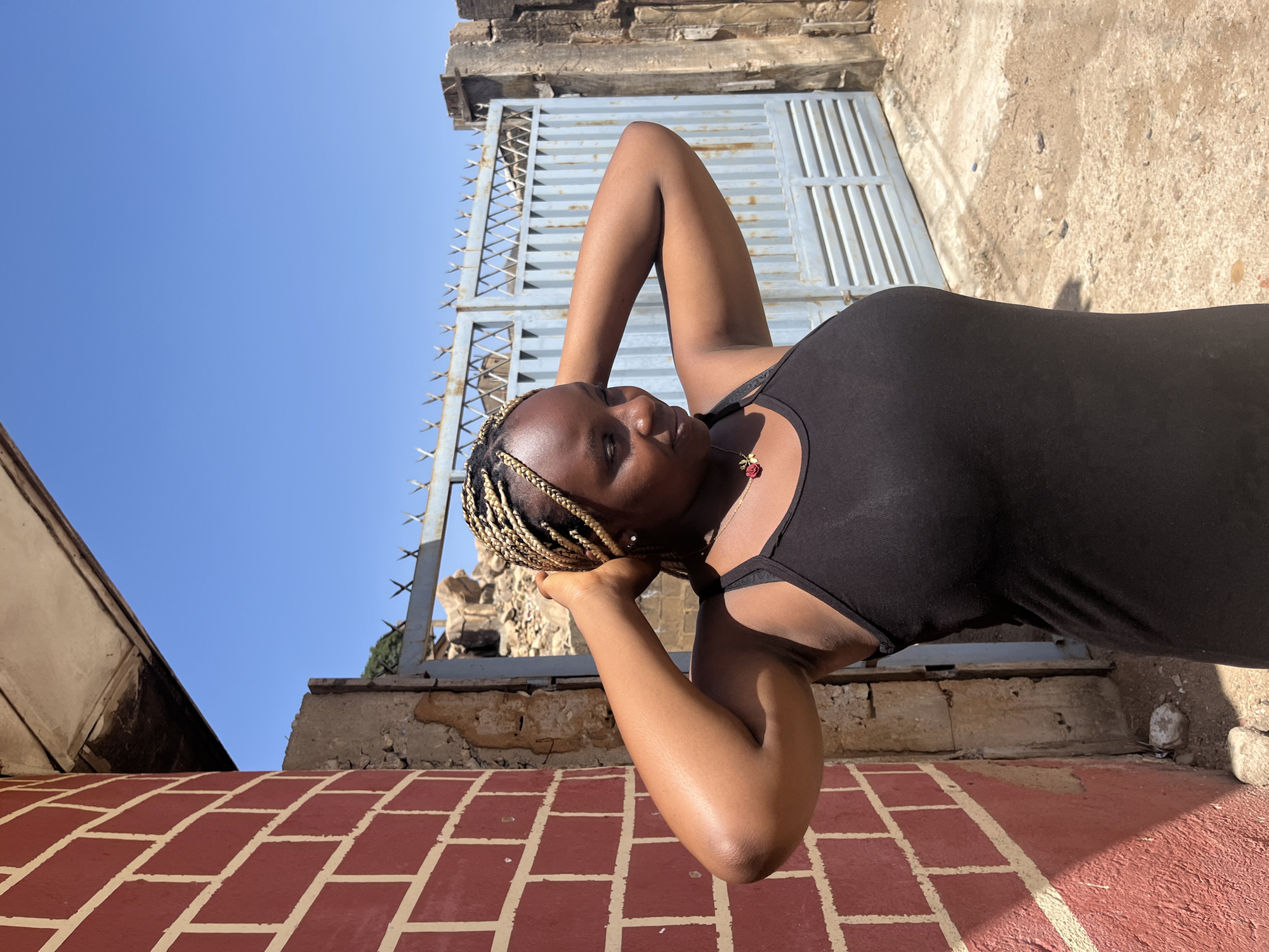 A woman with blonde-streaked braids wearing a black tank top is standing in front of a red-brick tile surface. Her arms are raised with her hands supporting her head. Behind her is a metal gate with spike-like points at the top. Behind the gate is a partially-destroyed concrete structure. The background is a clear blue sky. The location is Unknown, Unknown.