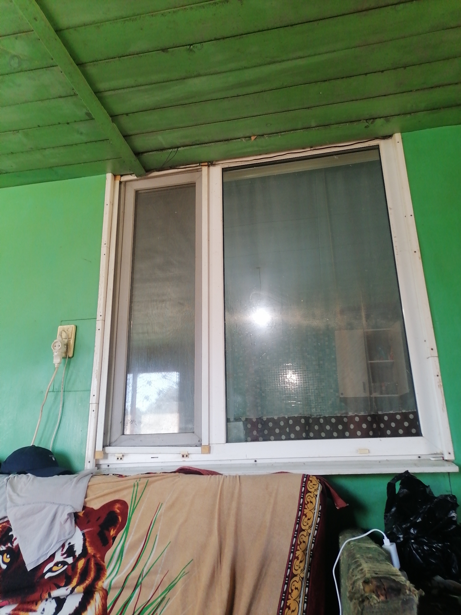 The image shows an interior view of a room, likely in Novopokrovka, Russia, featuring a large window with two panes. The left pane is covered by a screen, while the right pane is transparent, revealing a dimly lit interior with what appears to be furniture and possibly shelves. The walls of the room are painted a vibrant green, and the ceiling is made of green-painted wooden planks. To the left of the window, an electrical outlet with a plug and wire is visible. In the foreground, a blanket with a tiger print is draped over a surface, and a dark blue cap rests on top of some clothing. To the right, a black plastic bag and what appears to be a piece of furniture are visible. The overall impression is that of a functional but somewhat cluttered living space.
