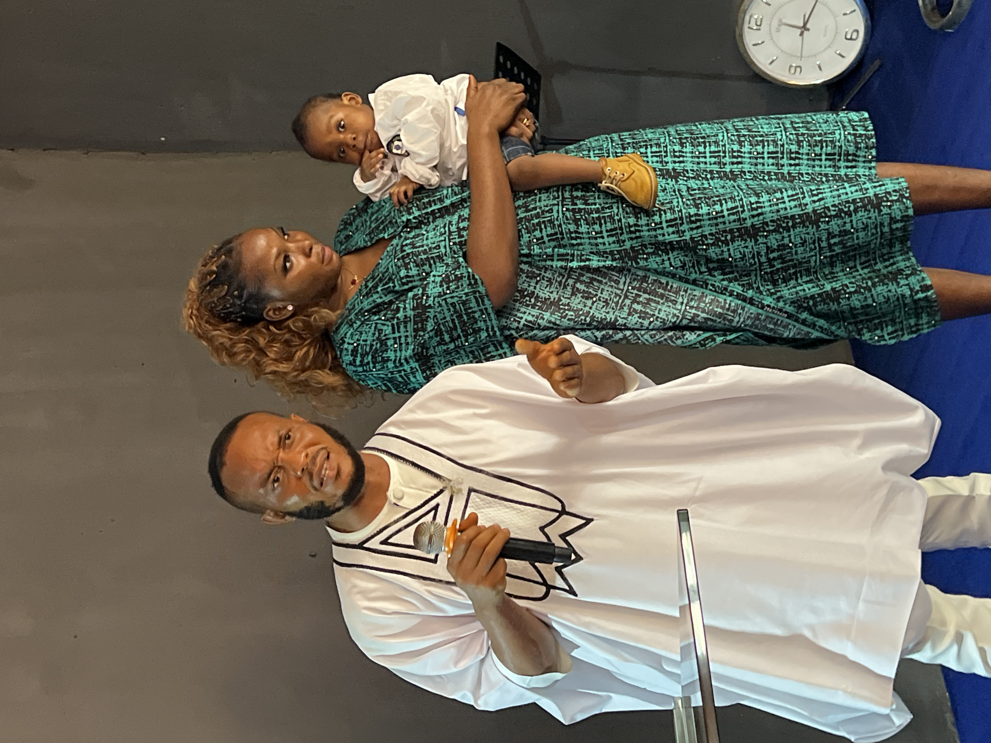 A man and a woman are posing for a photo with a baby in a room that appears to be a venue or church. The man, on the left, is wearing a white agbada with black trim and holding a microphone. He has a beard and a stern expression. The woman, on the right, is wearing a teal and black patterned dress and holding a baby. The baby is wearing a white shirt and blue shorts. The background is a plain gray wall. To the far right, a large analog clock is visible. This image was taken in the city of Nnewi, Nigeria.
