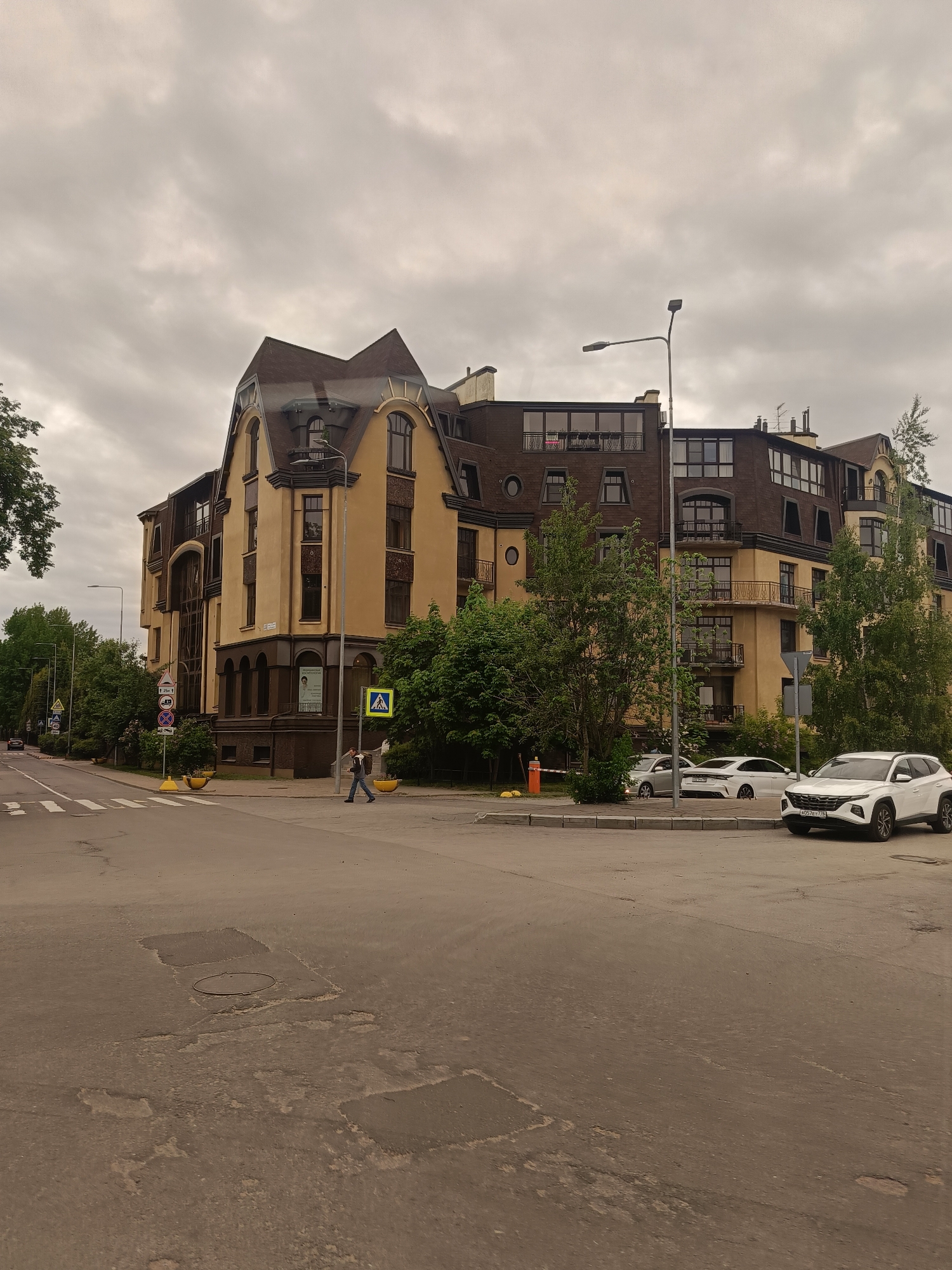 The image depicts a street scene in Saint Petersburg, Russia, under an overcast sky. A pedestrian is crossing a crosswalk at an intersection near a multi-story building with a distinctive architectural style, featuring a tan facade, brown roofing, and various window shapes. Several cars are parked along the side of the road. Street signs and greenery are visible, adding context to the urban setting. The road surface shows signs of wear and patches.
