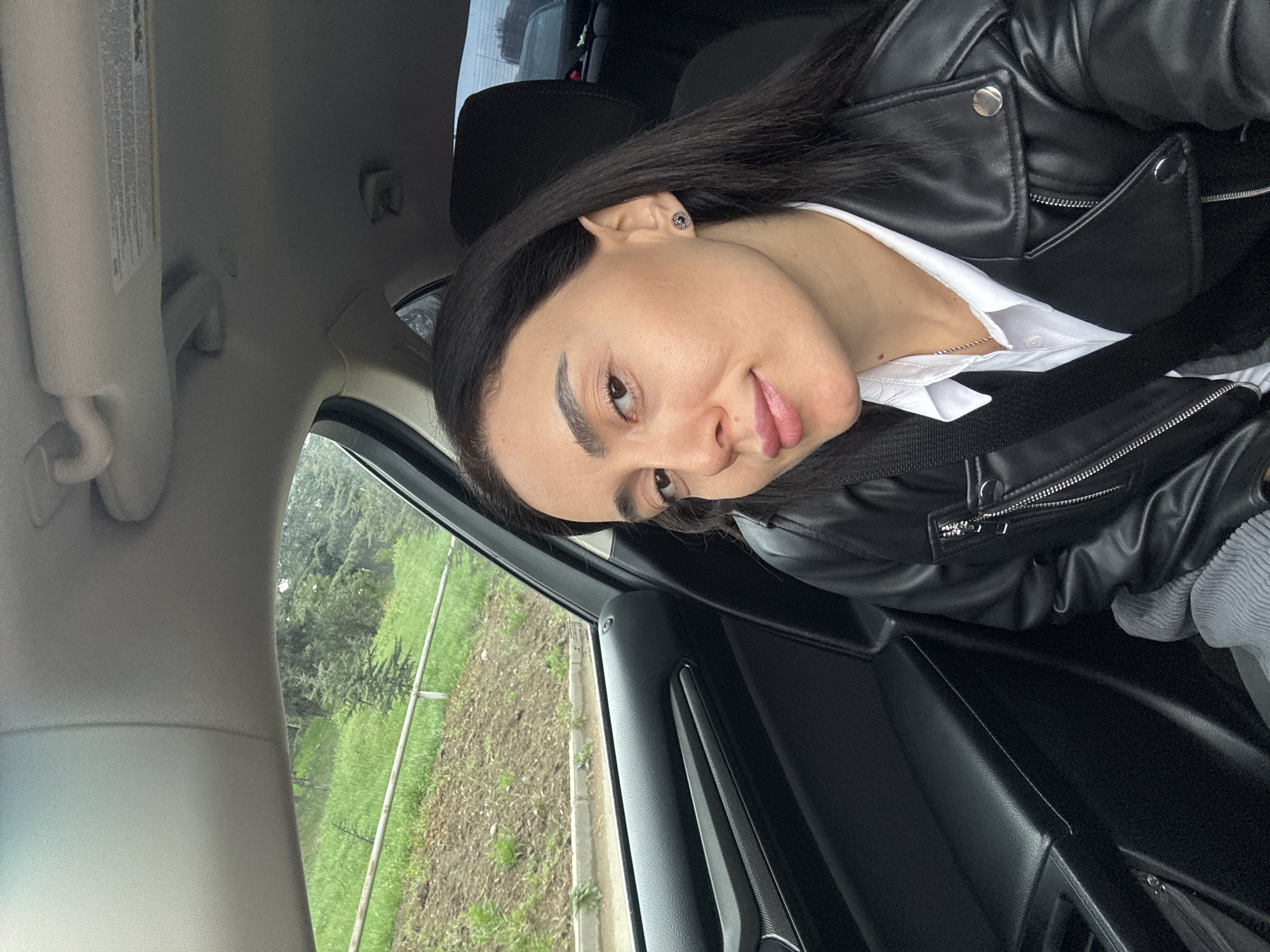A woman with dark hair, wearing a black leather jacket over a white collared shirt, is seated in the driver's seat of a car. She is wearing a seatbelt and looking at the camera. Outside the car window, there is a view of greenery, possibly a park or a roadside landscape. Due to the presence of greenery and the interior of the car, it's difficult to pinpoint a specific location in Tbilisi, Georgia, without further context. The car's interior is predominantly black.