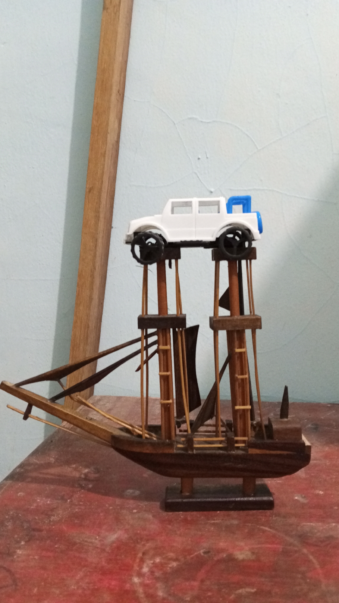 A small, brown, handcrafted wooden boat with a model white SUV on top of its elevated wooden pillars sits on a red surface. The boat features masts, rigging, and sails. The SUV model is white with black wheels and a blue detail. The background is a light blue wall with a wooden plank leaning against it. It cannot be determined if this is in Lhokseumawe, Indonesia from this image alone.