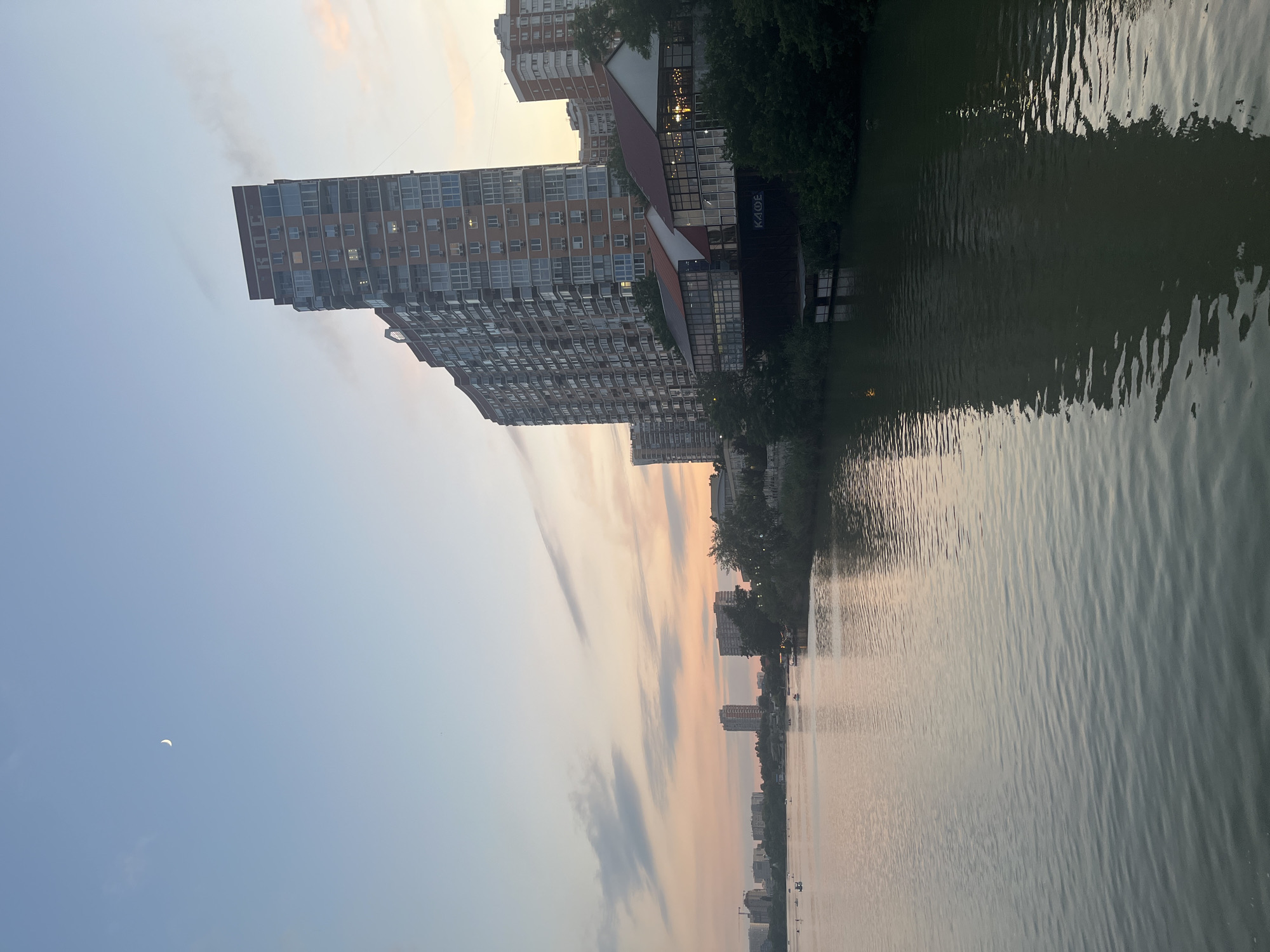 The image depicts a serene evening scene along the Kuban River in Krasnodar, Russia. On the left side of the frame, the tranquil waters of the river reflect the soft hues of the twilight sky. Several small boats are scattered across the river, with some people visible in them, enjoying the peaceful ambiance. The distant cityscape features a line of buildings, some of which appear to be under construction.
Dominating the right side of the image is a tall, modern residential building with a distinctive brick facade. Adjacent to this building, a low-rise structure with large glass windows and red-tiled roofs is visible, possibly a cafe or restaurant, with lights illuminated inside. The bank of the river is lined with dense green trees, creating a natural contrast to the urban architecture. The sky is a gradient of pale blue, transitioning into soft pinks and oranges near the horizon, indicating the sunset. A crescent moon is faintly visible in the upper left portion of the sky. The overall impression is one of urban tranquility at the close of day.