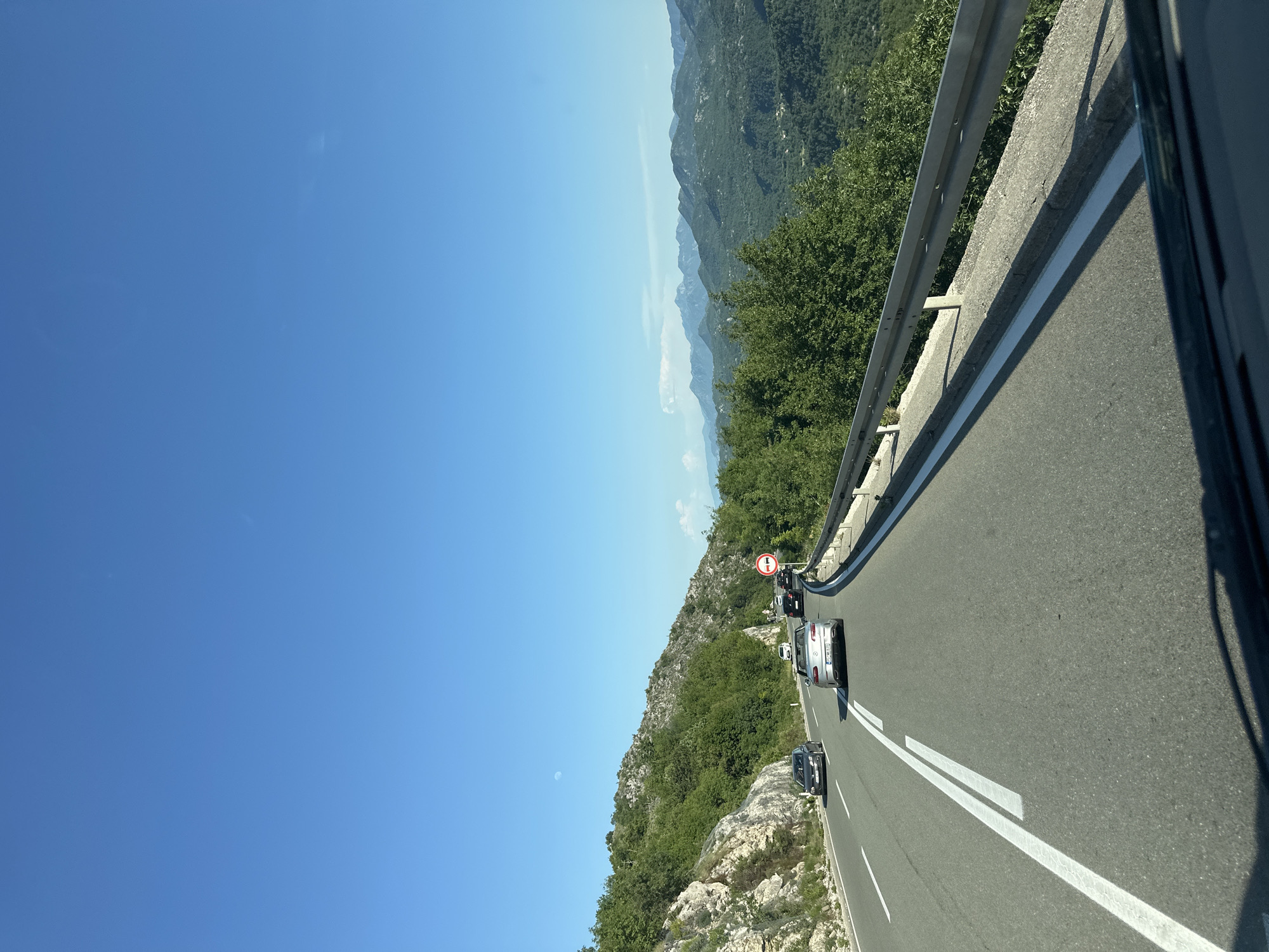 The image depicts a road in Dobrsko selo, Montenegro. The road is asphalt and has two white lines marking the lanes. There are several cars on the road, including a black car in the foreground and other vehicles further ahead. A guardrail runs along the side of the road, separating it from a steep, rocky slope covered in green vegetation. In the background, there are mountains covered in trees. The sky is clear and blue. There is a no-entry sign visible further along the road. The scene appears to be taken from inside a vehicle.