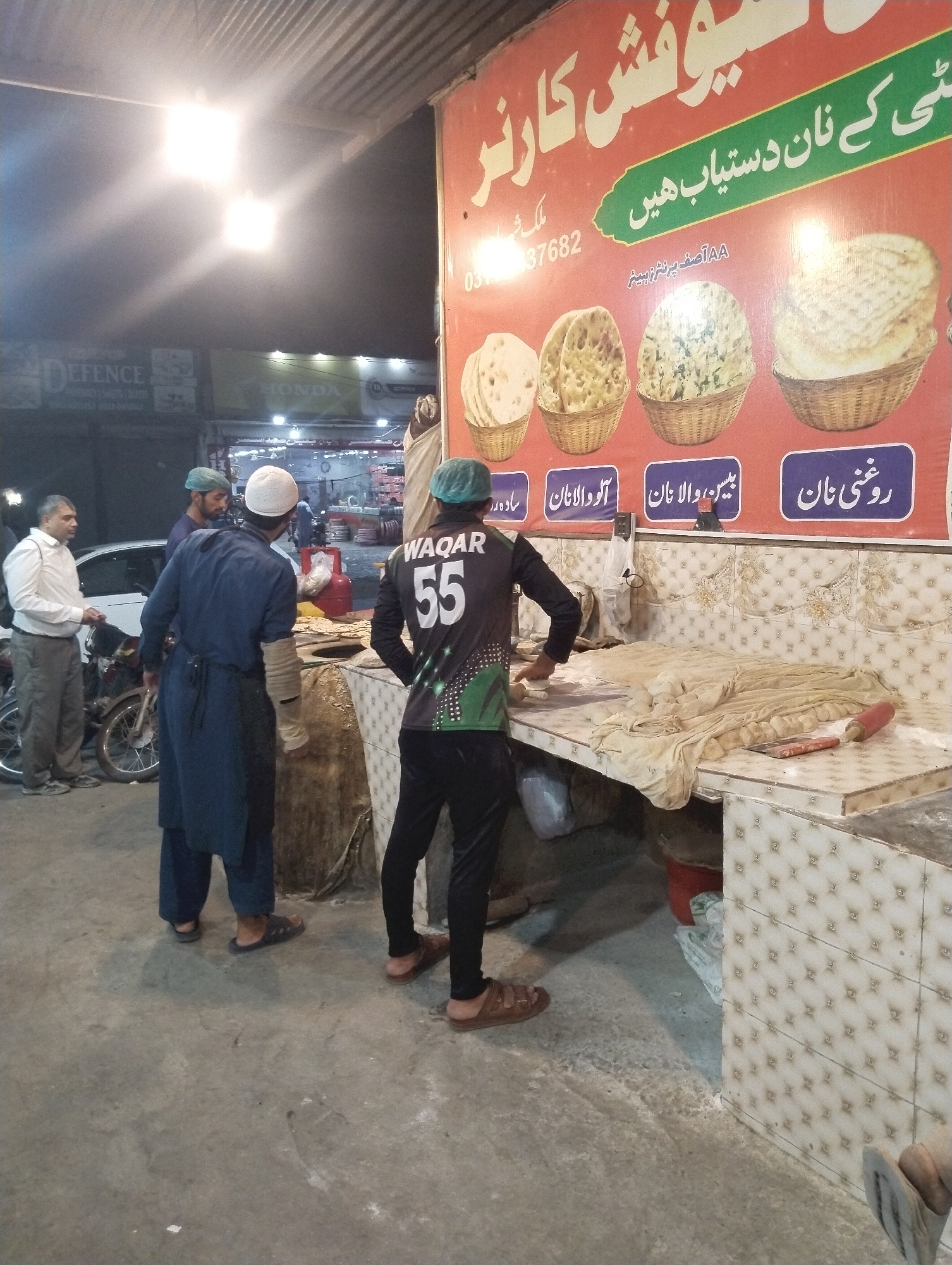The image depicts a nighttime street scene in Heir, Pakistan, centered around a bustling naan (flatbread) shop named "Yousaf Corner." A large, vibrant red and green signboard, written in Urdu, prominently displays the shop's name and advertises various types of naan available, including "Roghni Naan," "Besan Wala Naan," "Aloo Wala Naan," and "Sada Naan" (plain naan, partially visible). The sign also lists a contact number, 03137682.
Several men are present, engaged in activities related to the shop. On the far left, a man in a white shirt stands next to a parked motorcycle, possibly a customer. In the mid-ground, two men are near what appears to be a tandoor oven; one wears a white skullcap and a dark blue tunic, while the other has a green cap. To the right, a man wearing a black and green jersey with "WAQAR 55" printed on the back is actively rolling dough on a tiled counter, preparing naan. A rolling pin and a scraper are visible on the counter alongside the dough. A red gas cylinder can be seen beneath the counter.
The area is brightly illuminated by overhead fluorescent lights, creating some glare against the dark night sky. In the background, other commercial establishments with signs like "DEFENCE" and "HONDA" are faintly discernible, along with more motorcycles. The concrete ground shows signs of wear. The scene captures the active preparation and sale of fresh naan in a typical Pakistani street food setting.