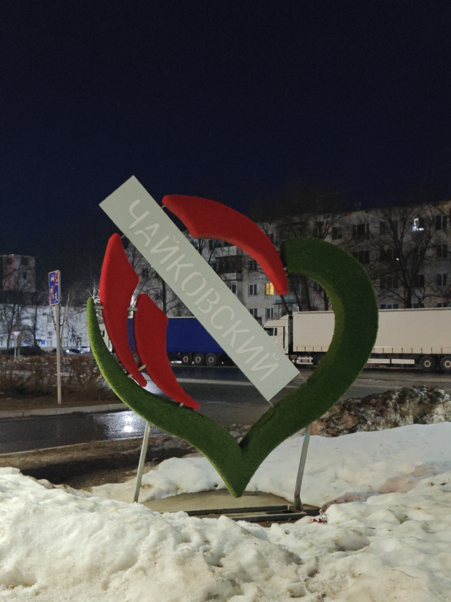 The image shows a sign in the city of Chaikovsky, Russia. The sign is composed of a green heart shape with red, flame-like accents and a diagonal white banner displaying the name "Чайковский" (Chaikovsky) in Cyrillic letters. The sign stands on metal supports and is surrounded by melting snow. In the background, a street with a truck and buildings is visible, under a dark, night sky. A street sign is also visible to the left of the sign.