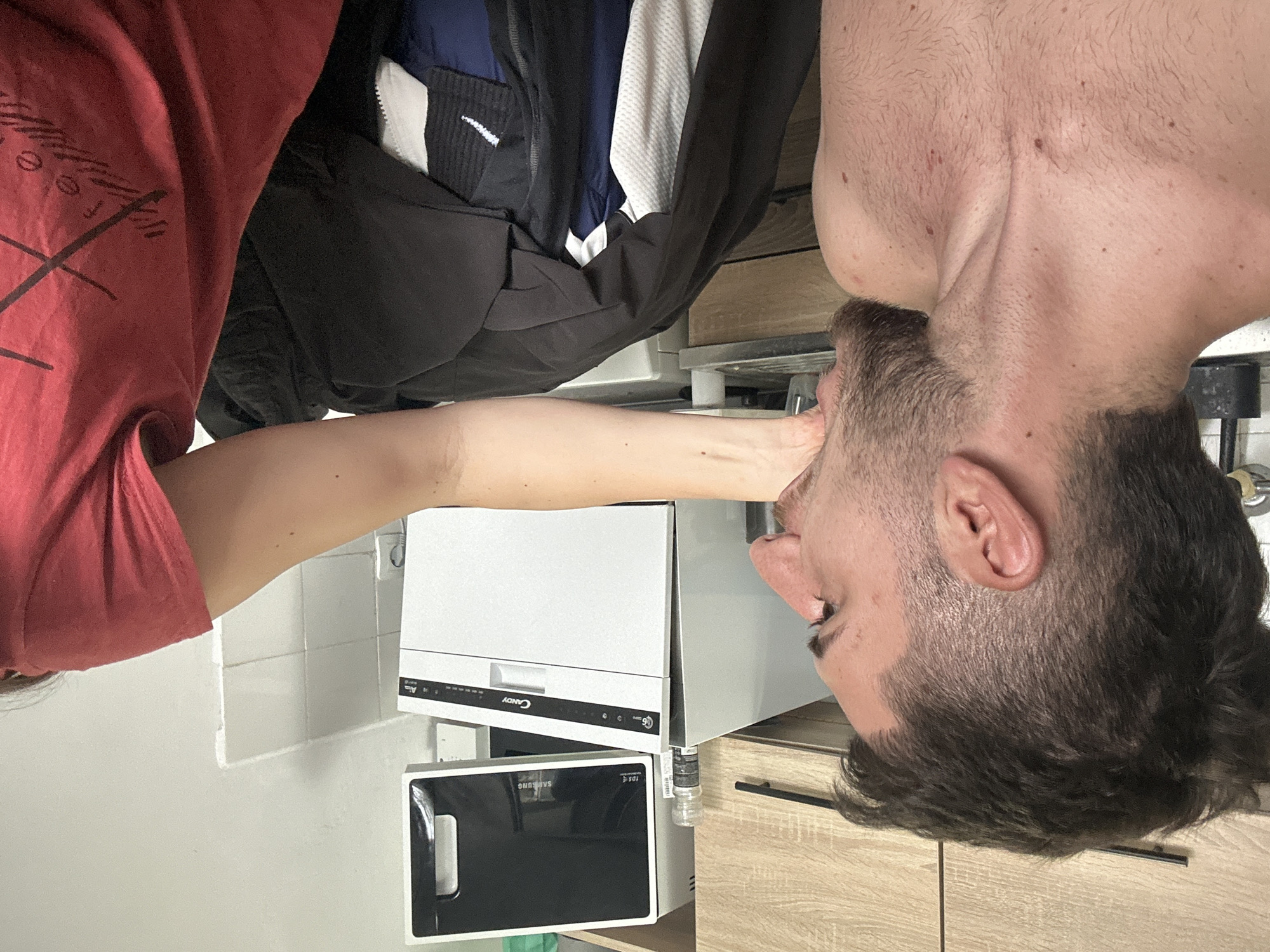 A man in a red t-shirt is positioned in front of kitchen appliances, including a Candy dishwasher and a Samsung microwave, in what appears to be a domestic setting. The photograph appears to be taken indoors, in Sofia, Bulgaria. The appliances are located along a tiled wall, and wooden cabinets are visible beneath the dishwasher. The man is reaching towards the appliances with his arm extended, his face closely oriented towards the appliances. Other clothing items (jacket and other apparels) are visible next to the man and positioned above the appliances.