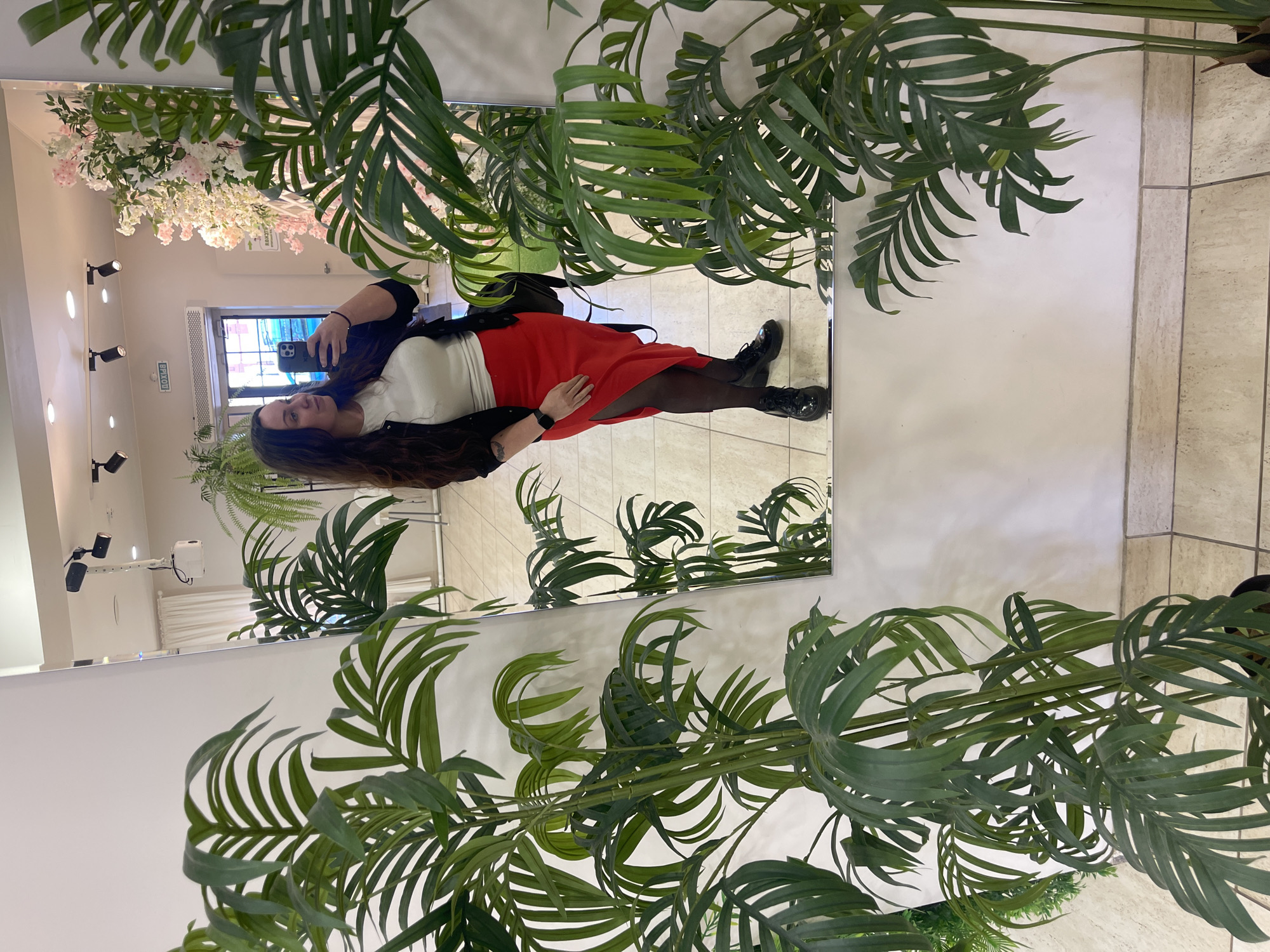 A woman is reflected in a mirror, taking a selfie with a smartphone. She is wearing a white top, a red skirt, a black jacket, black tights, and black boots. She has long, dark hair. The mirror is framed by artificial palm fronds and other greenery. In the background, the reflection shows a tiled floor, a doorway, floral decorations, and a series of spotlights. The sign above the door appears to be written in Cyrillic, which potentially indicates the location is in Russia. Based on the location hints given, the city is Osa.