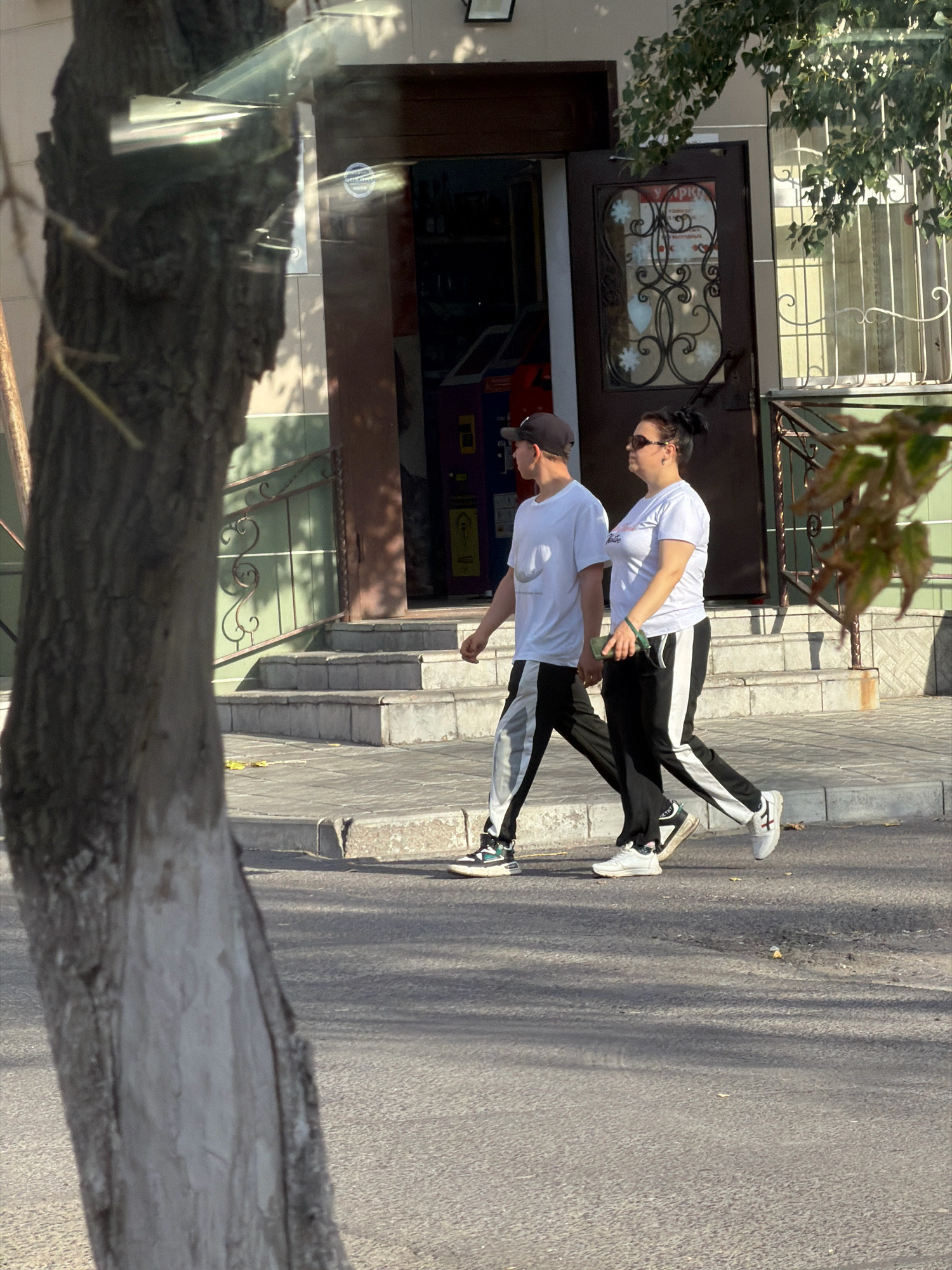 In Pavlodar, Kazakhstan, two individuals, a man and a woman, walk side-by-side on a paved sidewalk adjacent to a building with a dark wooden door and a barred window. The man, on the left, wears a dark baseball cap, a white t-shirt with a graphic, and black athletic pants with white stripes. The woman, on the right, wears sunglasses, a white t-shirt, and black athletic pants with white stripes. Both are wearing athletic shoes. The man's left hand is visible holding the woman's right hand. The woman is holding a green object, possibly a phone, in her right hand. A large tree trunk with textured bark is partially visible in the foreground, obscuring a portion of the scene. The background shows a building with steps leading up to an entrance. The lighting suggests it is daytime, with shadows cast across the sidewalk and street.
