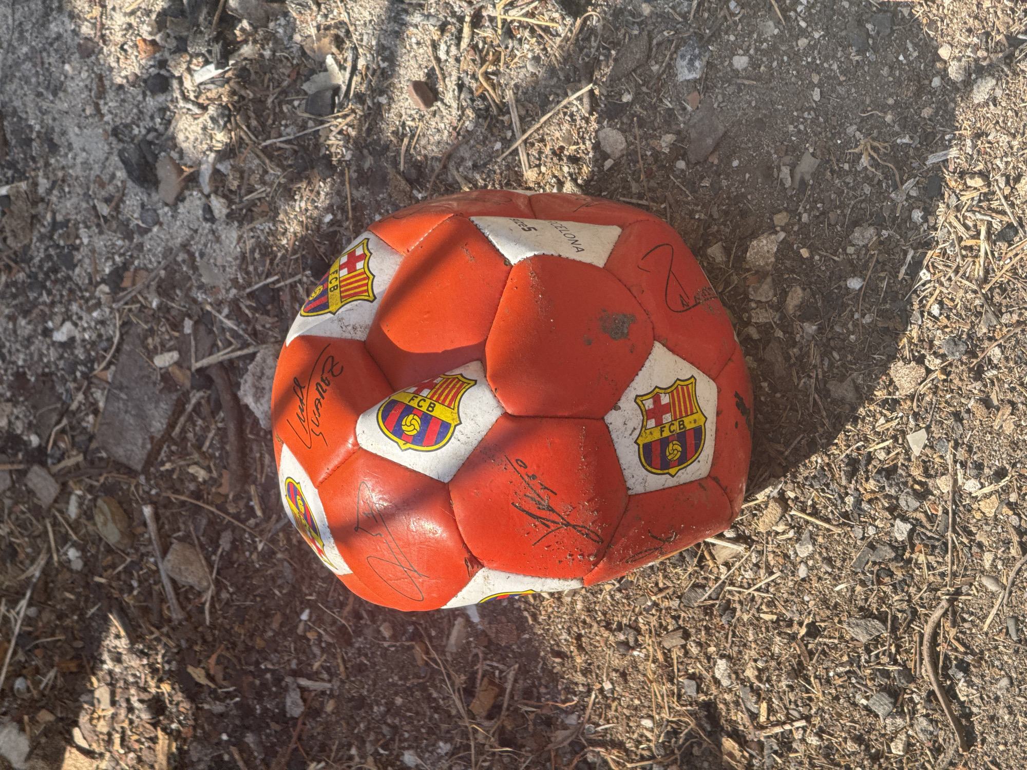 A red and white soccer ball lies on the ground in Zgrade, Montenegro. The ball is deflated and shows signs of wear and tear. It is covered in scuff marks and dirt. There are also several signatures on the ball, some of which are illegible. The background is a mix of dirt, rocks, and debris, suggesting an outdoor, possibly urban or semi-urban environment. The lighting indicates it is daytime, with shadows cast by an unseen object.