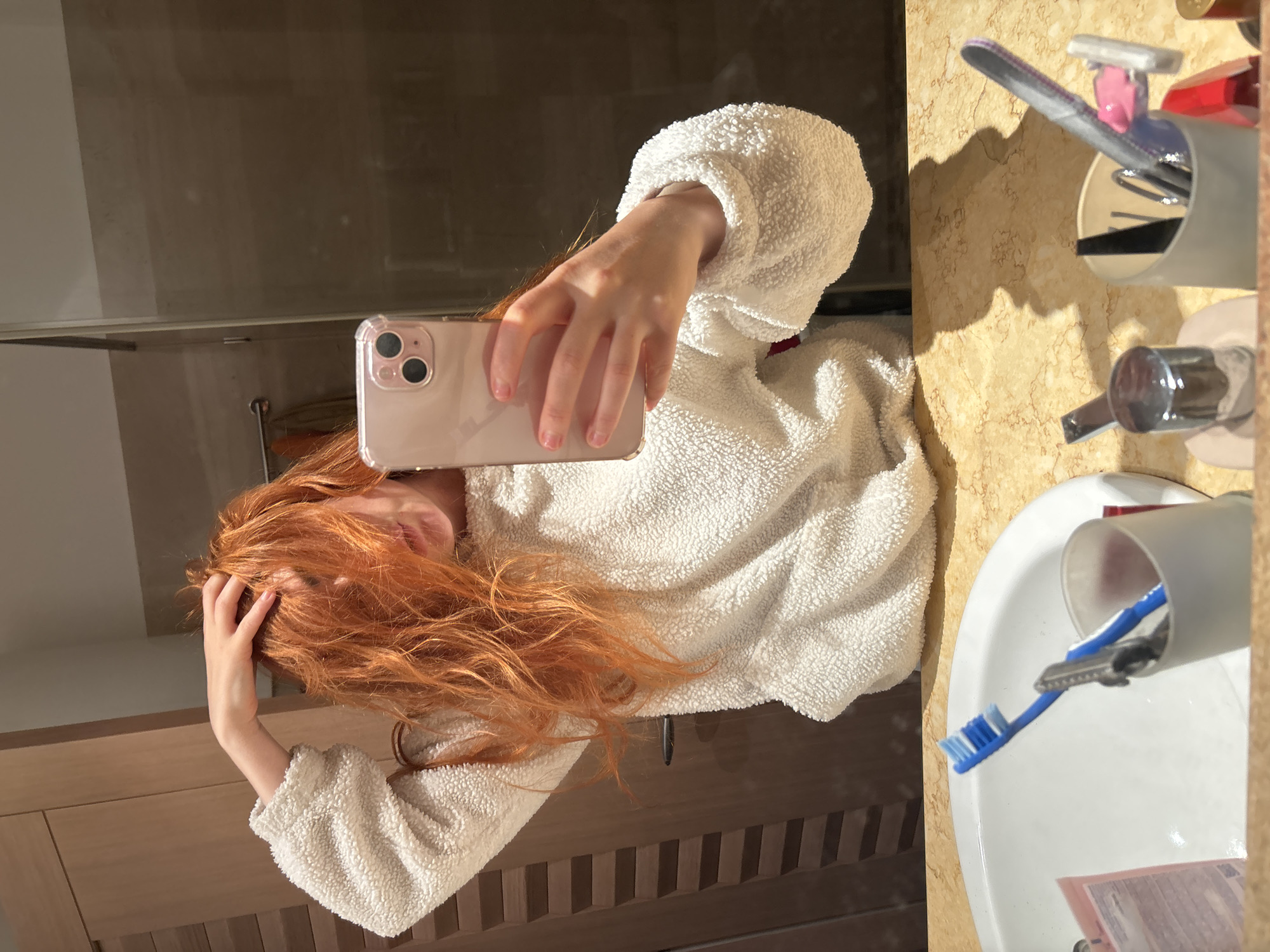 A woman with long, red hair is taking a selfie in a bathroom. She is wearing a fluffy, white sweater and holding a rose gold iPhone in her right hand. Her left hand is touching her hair. The bathroom features a sink, a wooden cabinet, and a marble countertop with a toothbrush holder containing toothbrushes and other items. The scene does not provide enough information to determine if the location is in the city of Budva, Montenegro.