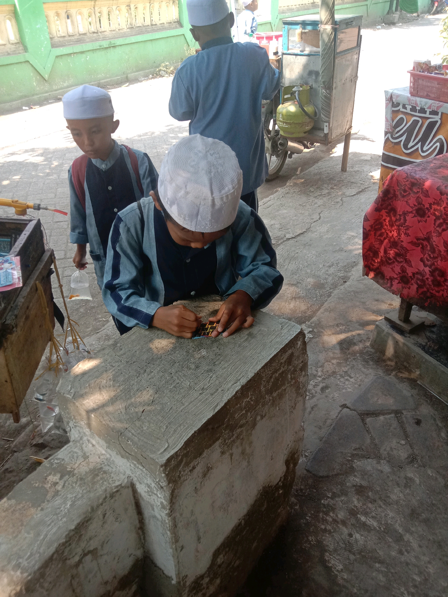 In Ngempit, Indonesia, a scene unfolds outdoors where two young boys, likely students due to their peci caps and matching uniforms, are engaged in activities near a roadside stall. The boy in the foreground, facing away from the camera, is intently focused on a small, colorful object he is manipulating with his hands. He wears a dark blue shirt with lighter blue accents and a peci cap. To his left and slightly behind him, another boy, also in similar attire and wearing a peci cap, is visible. He holds a clear plastic bag containing a small orange fish.
Behind the boys, a food cart with a gas cylinder and various items is parked. The cart appears to be made of metal and wood. To the right, a table is covered with a red floral patterned cloth, upon which a red basket and what appears to be a sign with text are placed. The ground is paved with concrete. In the background, a light green wall with decorative arches suggests the proximity of a building, possibly a mosque or school given the children's attire. The overall environment suggests a typical street scene in a village or town, likely during daytime.